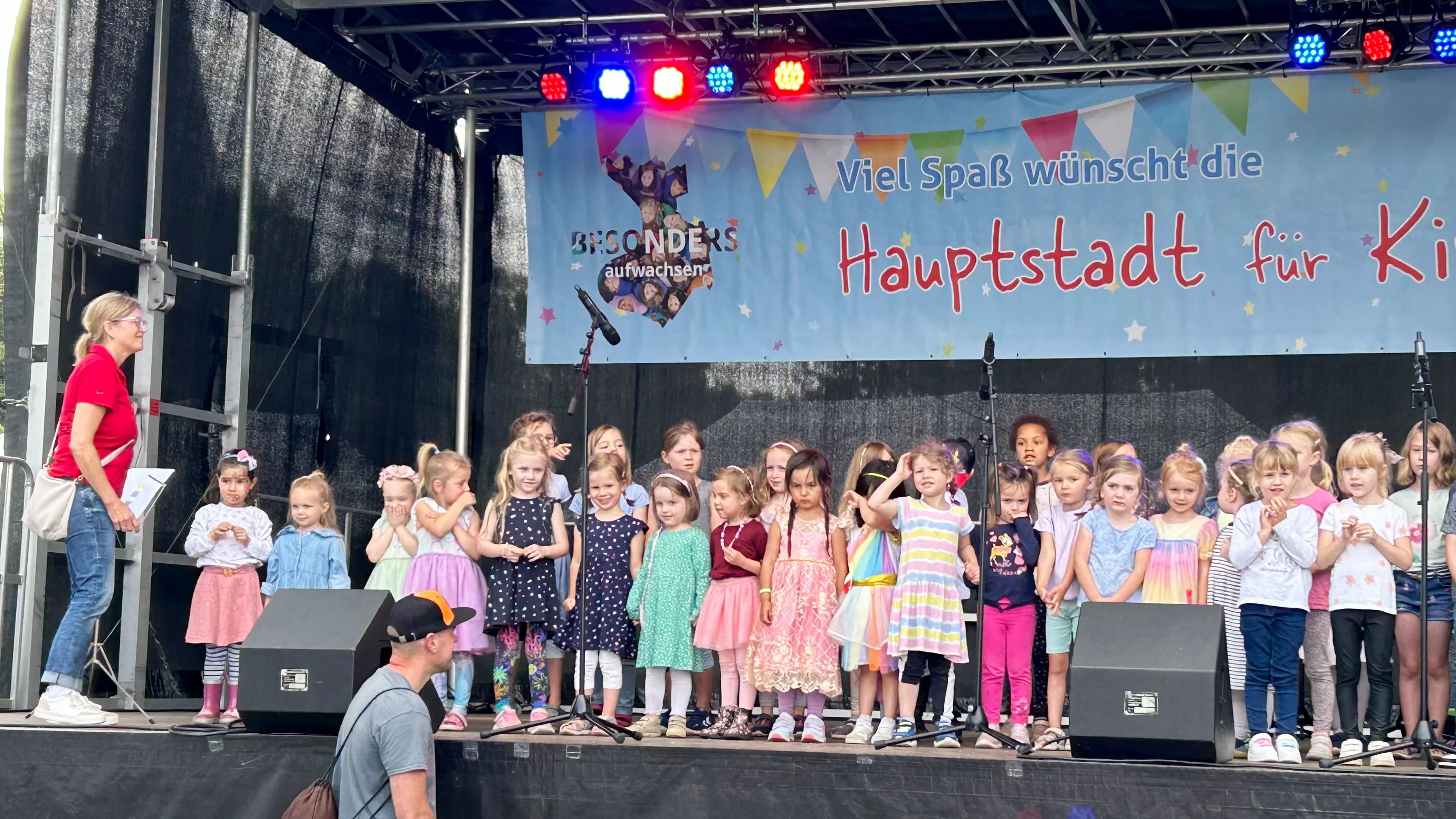 Auf dem Foto ist eine Gruppe von Kindern auf einer Bühne zu sehen, begleitet von einigen Erwachsenen. Die Kinder nehmen an einer Veranstaltung oder Aufführung teil und tragen bunte Kleidung. Und die Kulisse ist eine Bühne mit einem Hintergrund, auf dem Text zu sehen ist. Der Text auf dem Hintergrund ist auf Deutsch und lautet: “Viel Spaß wünscht die Hauptstadt für Kinder.”