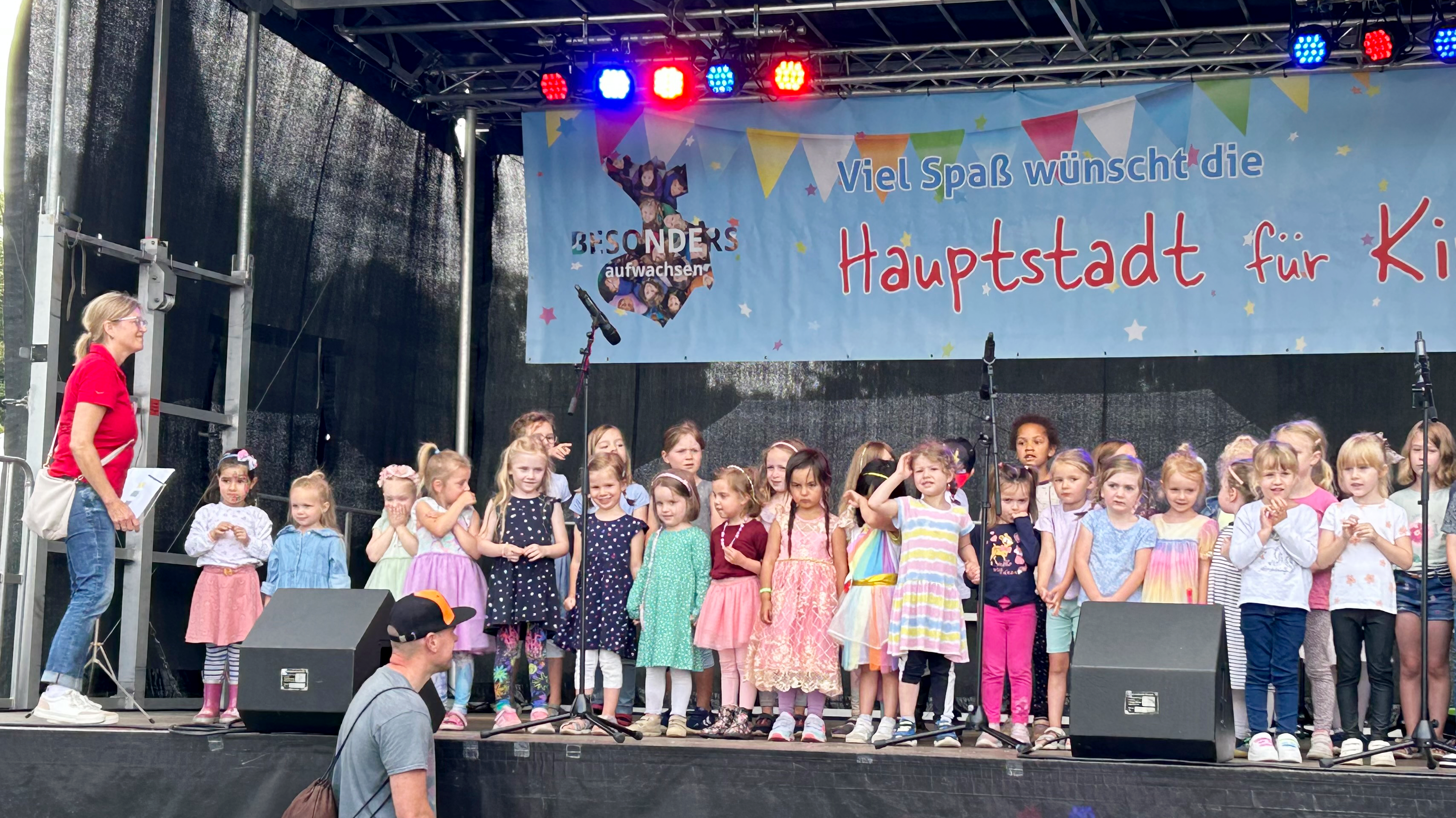 Auf dem Foto ist eine Gruppe von Kindern auf einer Bühne zu sehen, begleitet von einigen Erwachsenen. Die Kinder nehmen an einer Veranstaltung oder Aufführung teil und tragen bunte Kleidung. Und die Kulisse ist eine Bühne mit einem Hintergrund, auf dem Text zu sehen ist. Der Text auf dem Hintergrund ist auf Deutsch und lautet: “Viel Spaß wünscht die Hauptstadt für Kinder.”