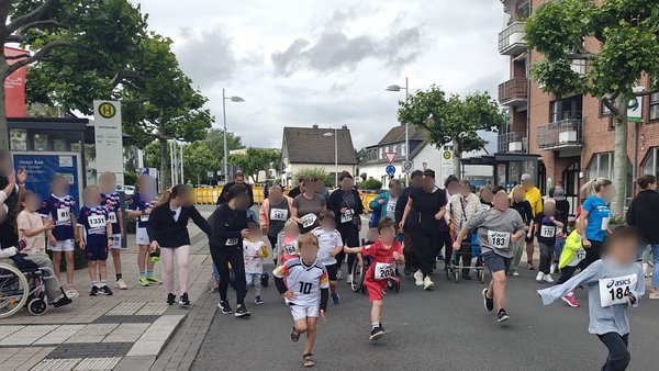  Auf dem Bild ist ein Kinder- und Familienlauf in einer Innenstadt-Straße zu sehen, vermutlich im Rahmen einer lokalen Lauf- oder Stadtveranstaltung. Szene und Personen Mehrere Kinder mit Startnummern rennen auf einer für den Verkehr gesperrten Straße los, einige in Sportkleidung, andere in Alltagskleidung. Am Rand der Straße stehen und sitzen zahlreiche Erwachsene, teilweise mit Kinderwagen oder Rollstühlen, die zuschauen, anfeuern oder die Kinder begleiten. Umgebung Die Szene spielt an einer Bushaltestelle vor mehrstöckigen Wohn- und Geschäftshäusern, rechts im Bild sind Ladenfassaden mit auffälligen Logos und ein großes Informationsplakat zu sehen. Der Boden ist eine typische Stadtstraße mit Gehwegen, Bordstein und taktilen Platten, und der Himmel ist bewölkt, was auf eher kühles, aber trockenes Wetter schließen lässt.