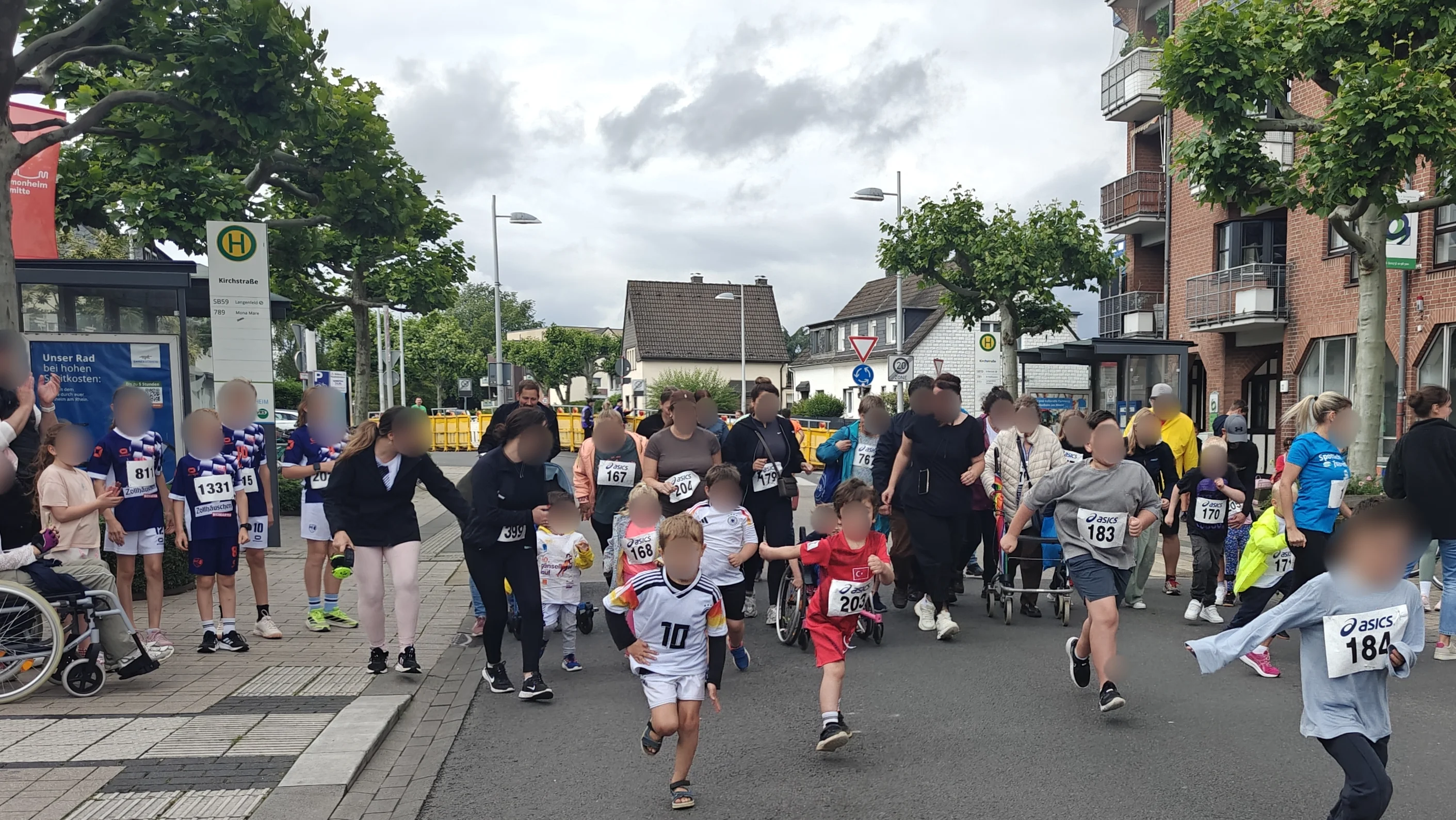  Auf dem Bild ist ein Kinder- und Familienlauf in einer Innenstadt-Straße zu sehen, vermutlich im Rahmen einer lokalen Lauf- oder Stadtveranstaltung. Szene und Personen Mehrere Kinder mit Startnummern rennen auf einer für den Verkehr gesperrten Straße los, einige in Sportkleidung, andere in Alltagskleidung. Am Rand der Straße stehen und sitzen zahlreiche Erwachsene, teilweise mit Kinderwagen oder Rollstühlen, die zuschauen, anfeuern oder die Kinder begleiten. Umgebung Die Szene spielt an einer Bushaltestelle vor mehrstöckigen Wohn- und Geschäftshäusern, rechts im Bild sind Ladenfassaden mit auffälligen Logos und ein großes Informationsplakat zu sehen. Der Boden ist eine typische Stadtstraße mit Gehwegen, Bordstein und taktilen Platten, und der Himmel ist bewölkt, was auf eher kühles, aber trockenes Wetter schließen lässt.