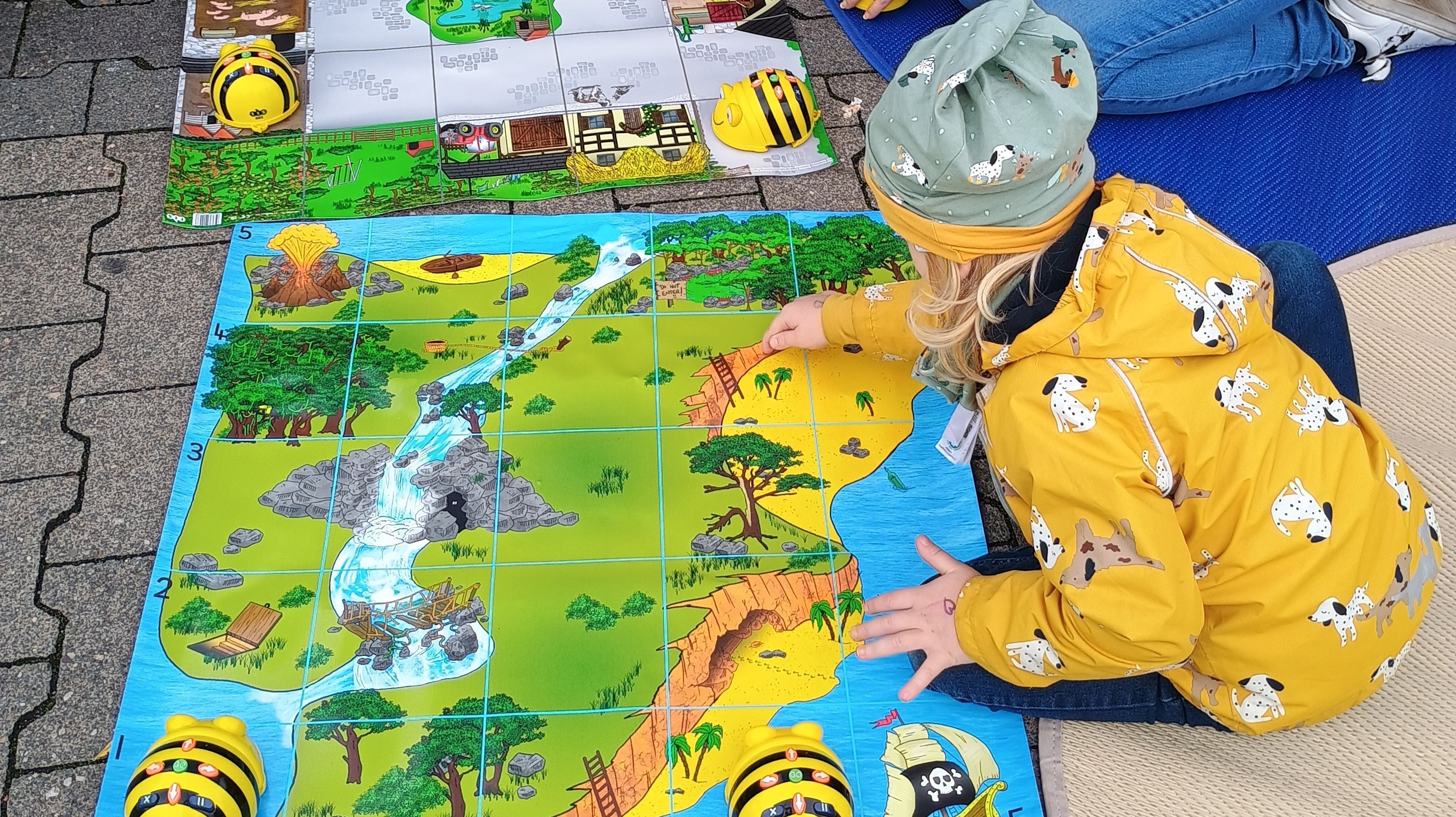  Auf dem Foto ist eine kleine Gruppe von Kindern und einer erwachsenen Person zu sehen, die an einer Programmier- oder Lernaktivität teilnehmen, die im Freien stattfindet. Hier sind die Details: Aktivität: Sie sitzen auf dem Boden um zwei bunte Matten herum. Die Matten sind in Quadrate unterteilt (ähnlich einem Raster) und zeigen thematische Landschaften (eine Dschungel-/Flusslandschaft und eine Parklandschaft). Geräte: Auf den Matten befinden sich mehrere gelb-schwarze, bienenförmige Roboter (sogenannte Bee-Bots oder vergleichbare Bodenroboter). Diese werden in der frühkindlichen Bildung häufig zur spielerischen Einführung in die Programmierung genutzt. Die Kinder scheinen sie über die Matten zu navigieren. Personen: Mehrere Kinder sind in warme Kleidung (Jacken und Mützen) gekleidet. Eine erwachsene Person (vermutlich eine Betreuungsperson oder Elternteil) kniet bei den Kindern und unterstützt sie bei der Aktivität. Umgebung: Die Szene findet auf einem gepflasterten Außenbereich statt, und es liegen weiche Matten unter den Spielenden.