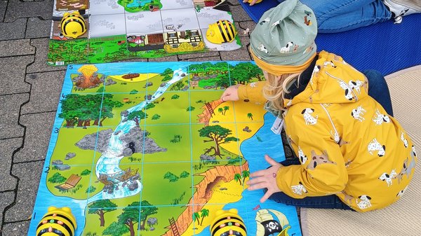  Auf dem Foto ist eine kleine Gruppe von Kindern und einer erwachsenen Person zu sehen, die an einer Programmier- oder Lernaktivität teilnehmen, die im Freien stattfindet. Hier sind die Details: Aktivität: Sie sitzen auf dem Boden um zwei bunte Matten herum. Die Matten sind in Quadrate unterteilt (ähnlich einem Raster) und zeigen thematische Landschaften (eine Dschungel-/Flusslandschaft und eine Parklandschaft). Geräte: Auf den Matten befinden sich mehrere gelb-schwarze, bienenförmige Roboter (sogenannte Bee-Bots oder vergleichbare Bodenroboter). Diese werden in der frühkindlichen Bildung häufig zur spielerischen Einführung in die Programmierung genutzt. Die Kinder scheinen sie über die Matten zu navigieren. Personen: Mehrere Kinder sind in warme Kleidung (Jacken und Mützen) gekleidet. Eine erwachsene Person (vermutlich eine Betreuungsperson oder Elternteil) kniet bei den Kindern und unterstützt sie bei der Aktivität. Umgebung: Die Szene findet auf einem gepflasterten Außenbereich statt, und es liegen weiche Matten unter den Spielenden.