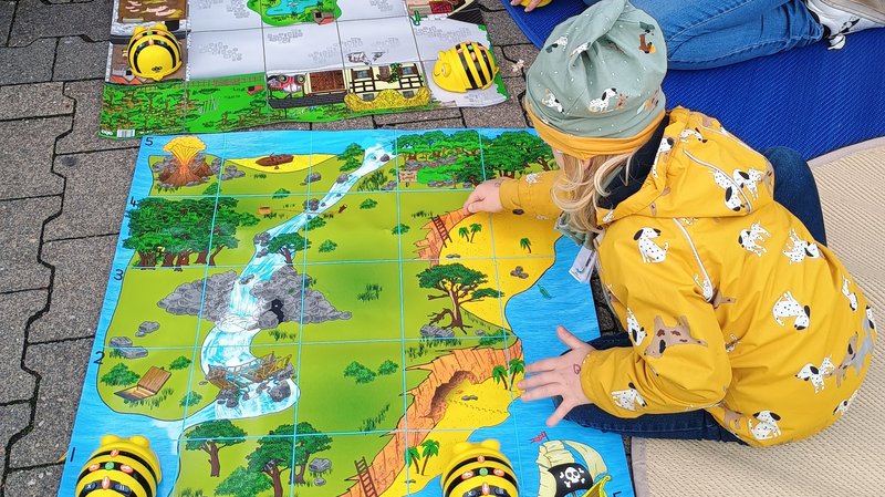  Auf dem Foto ist eine kleine Gruppe von Kindern und einer erwachsenen Person zu sehen, die an einer Programmier- oder Lernaktivität teilnehmen, die im Freien stattfindet. Hier sind die Details: Aktivität: Sie sitzen auf dem Boden um zwei bunte Matten herum. Die Matten sind in Quadrate unterteilt (ähnlich einem Raster) und zeigen thematische Landschaften (eine Dschungel-/Flusslandschaft und eine Parklandschaft). Geräte: Auf den Matten befinden sich mehrere gelb-schwarze, bienenförmige Roboter (sogenannte Bee-Bots oder vergleichbare Bodenroboter). Diese werden in der frühkindlichen Bildung häufig zur spielerischen Einführung in die Programmierung genutzt. Die Kinder scheinen sie über die Matten zu navigieren. Personen: Mehrere Kinder sind in warme Kleidung (Jacken und Mützen) gekleidet. Eine erwachsene Person (vermutlich eine Betreuungsperson oder Elternteil) kniet bei den Kindern und unterstützt sie bei der Aktivität. Umgebung: Die Szene findet auf einem gepflasterten Außenbereich statt, und es liegen weiche Matten unter den Spielenden.