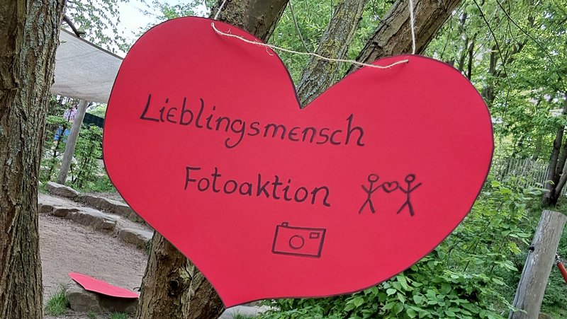 Auf dem Bild hängt ein großes rotes Herz‑Schild in einem Baum in einer grünen Außenanlage. Auf dem Herz steht in schwarzer Schrift „Lieblingsmensch Fotoaktion“, dazu sind zwei Strichmännchen mit einem Herz darüber und ein kleines Kamerasymbol gezeichnet.