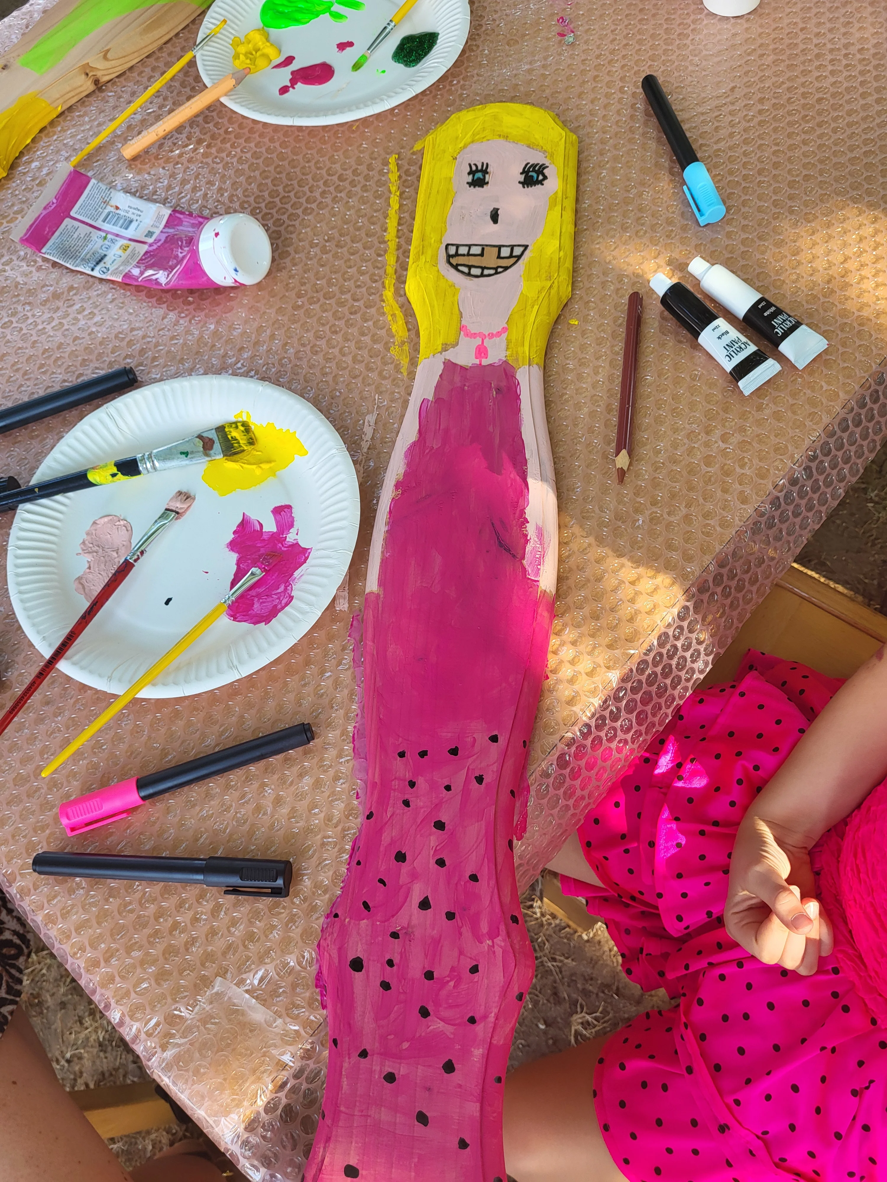 Auf dem Bild sieht man eine Bastel- und Malsituation. In der Mitte liegt ein längliches Holzbrett, auf das eine Figur mit gelben Haaren und pinkem Kleid gemalt ist; die Figur hat ein großes lächelndes Gesicht mit sichtbaren Zähnen und einem rosa Halskettendetail. Drumherum liegen Papierteller mit bunter Farbe (vor allem Pink, Gelb, Grün), Pinsel, Filzstifte und Farbtuben auf einer mit Noppenfolie abgedeckten Tischplatte. Rechts unten ist ein Kind im pink-schwarz gepunkteten Kleid oder Rock zu sehen, dessen Kleidung farblich gut zu der bemalten Figur passt.