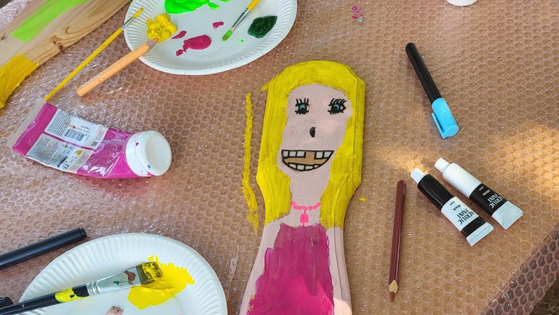 Auf dem Bild sieht man eine Bastel- und Malsituation. In der Mitte liegt ein längliches Holzbrett, auf das eine Figur mit gelben Haaren und pinkem Kleid gemalt ist; die Figur hat ein großes lächelndes Gesicht mit sichtbaren Zähnen und einem rosa Halskettendetail. Drumherum liegen Papierteller mit bunter Farbe (vor allem Pink, Gelb, Grün), Pinsel, Filzstifte und Farbtuben auf einer mit Noppenfolie abgedeckten Tischplatte. Rechts unten ist ein Kind im pink-schwarz gepunkteten Kleid oder Rock zu sehen, dessen Kleidung farblich gut zu der bemalten Figur passt.