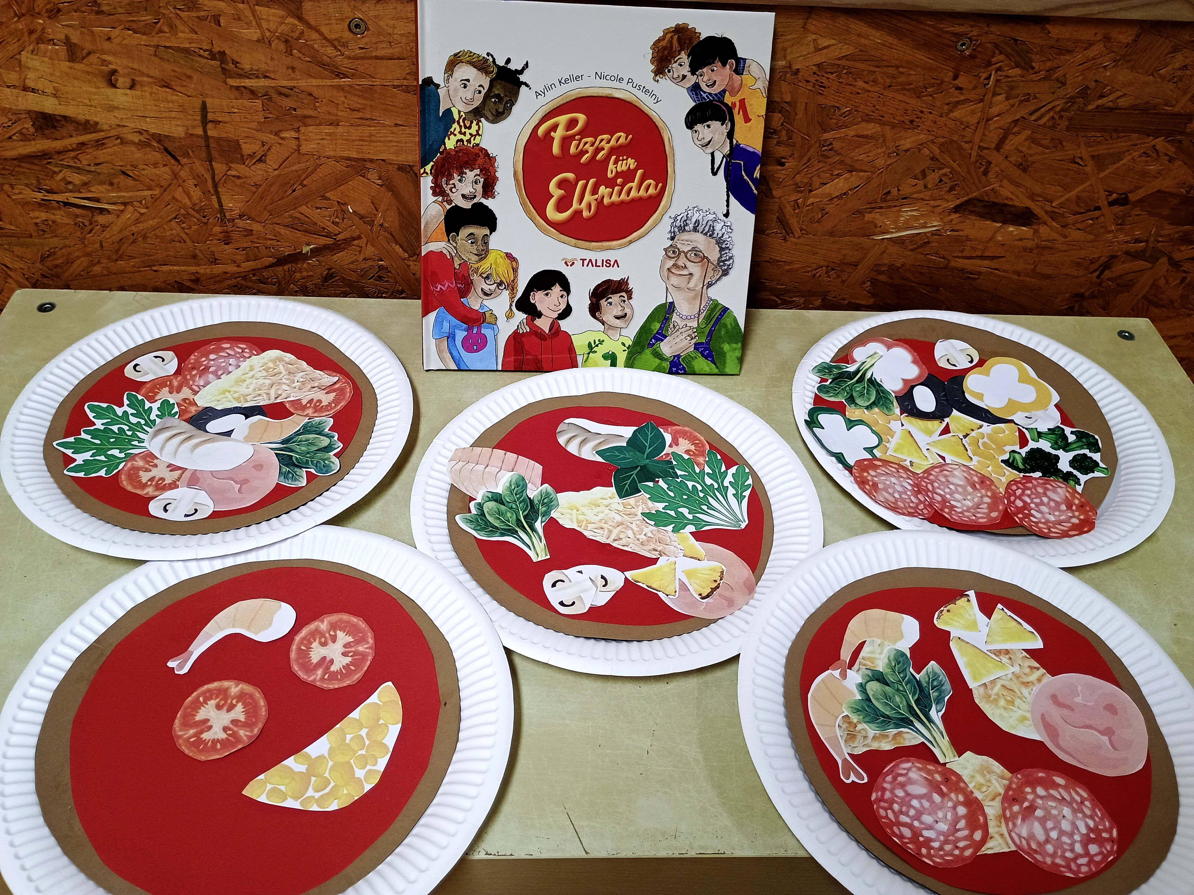 Auf dem Foto ist ein Bastelangebot mit Papier‑Pizzen zu sehen. Vorne liegen fünf auf Papptellern gestaltete „Pizzen“, die aus verschiedenfarbigem Tonpapier und ausgeschnittenen Belägen wie Salami, Käse, Pilzen, Tomaten, Spinat und Mais bestehen. Hinter den Tellern steht ein Kinderbuch mit dem Titel „Pizza für Elfrida“, dessen Cover mehrere gezeichnete Personen zeigt.