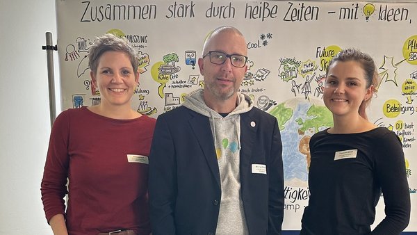 Auf dem Bild sind drei Personen – eine Frau links, ein Mann in der Mitte und eine Frau rechts – zu sehen, die vor einer Whiteboard- oder Plakatwand stehen. Die Wand hinter ihnen enthält eine Überschrift, die teilweise lesbar ist: "Zusammen stark durch heiße Zeiten - mit Ideen". Darunter ist eine bunte, skizzenhafte Weltkarte und verschiedene Piktogramme und Notizen in verschiedenen Farben gezeichnet, die sich wahrscheinlich auf die Überschrift beziehen. Alle drei Personen tragen Namensschilder. Sie schauen in die Kamera und lächeln leicht.