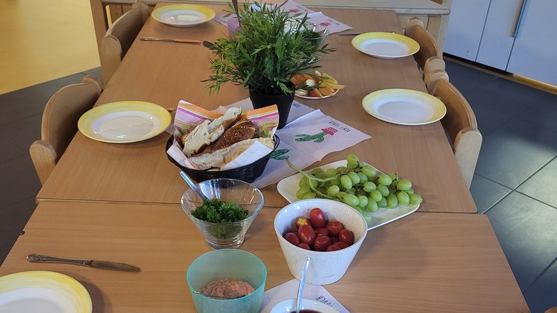 Auf dem Bild ist ein gedeckter Tisch in einem Gemeinschafts- oder Gruppenraum zu sehen. Darauf stehen unter anderem: Teller, Besteck und bunte Servietten Ein Korb mit belegten Broten Weintrauben und Kirschtomaten Aufgeschnittene Gurken Verschiedene Aufstriche oder Dips Eine Schale mit Kräutern Eine Topfpflanze als Tischdekoration