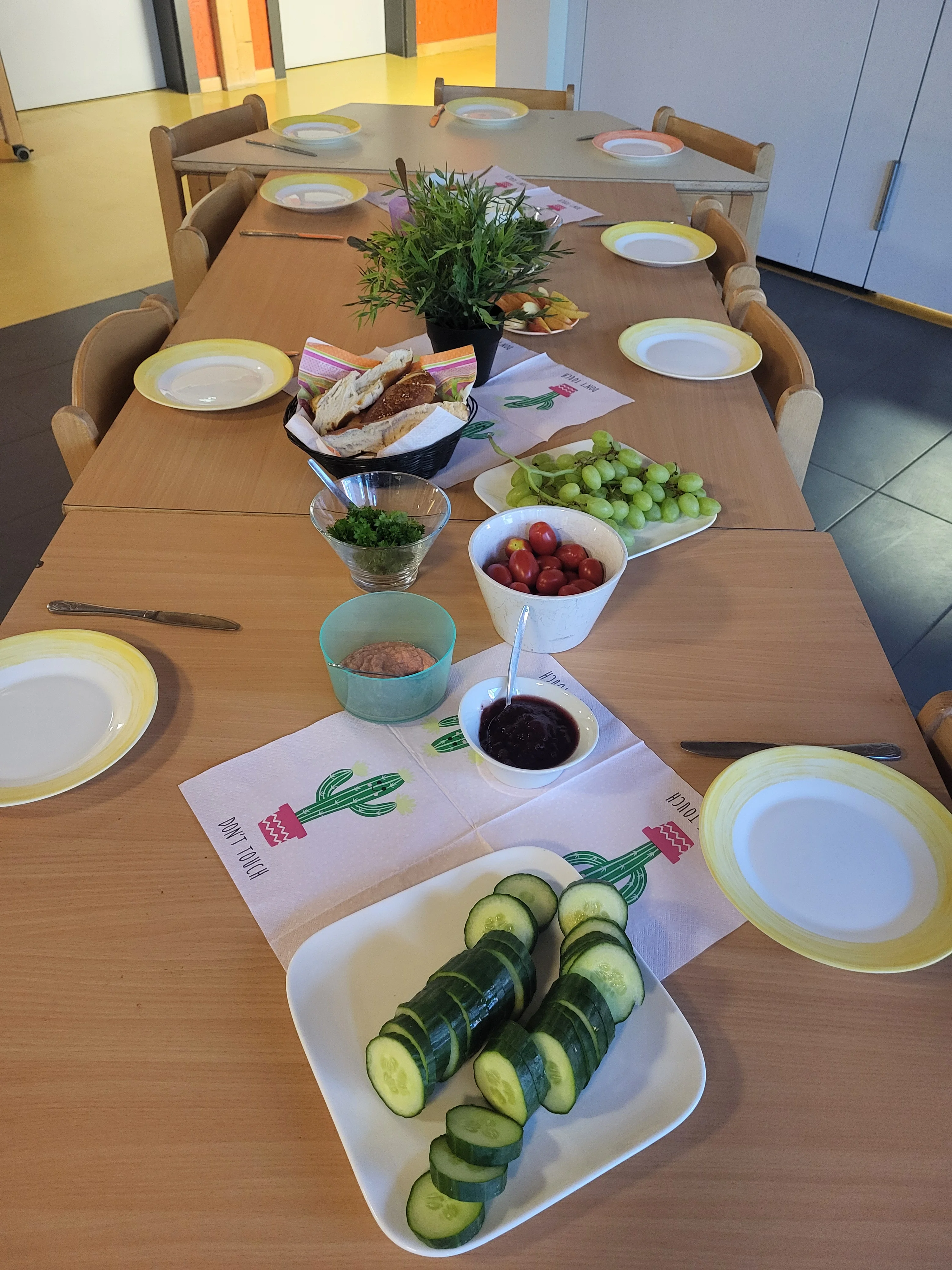 Auf dem Bild ist ein gedeckter Tisch in einem Gemeinschafts- oder Gruppenraum zu sehen. Darauf stehen unter anderem: Teller, Besteck und bunte Servietten Ein Korb mit belegten Broten Weintrauben und Kirschtomaten Aufgeschnittene Gurken Verschiedene Aufstriche oder Dips Eine Schale mit Kräutern Eine Topfpflanze als Tischdekoration