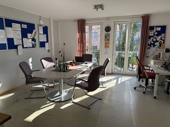 Das Bild zeigt ein Besprechungszimmer mit vier Stühlen um einen runden Tisch. Auf dem Tisch befinden sich mehrere Flaschen, eine Tasse, ein Laptop und einige Papiere. Im Hintergrund sind zwei Pinnwände mit verschiedenen Notizen und Dokumenten zu sehen. Es gibt auch einen Schreibtisch mit einem Computer auf der rechten Seite des Raumes. Die Fenster lassen viel Tageslicht herein, und draußen sind Bäume und ein Gebäude sichtbar.