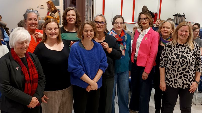 Auf dem Foto ist eine Gruppe von Menschen in einem Veranstaltungsraum zu sehen, die dicht beieinandersteht und in Richtung Kamera schaut. Im Hintergrund sitzen weitere Personen, außerdem sind Tische, Stühle, eine Garderobe, eine Uhr, Türen und ein Beamer an der Decke zu erkennen.