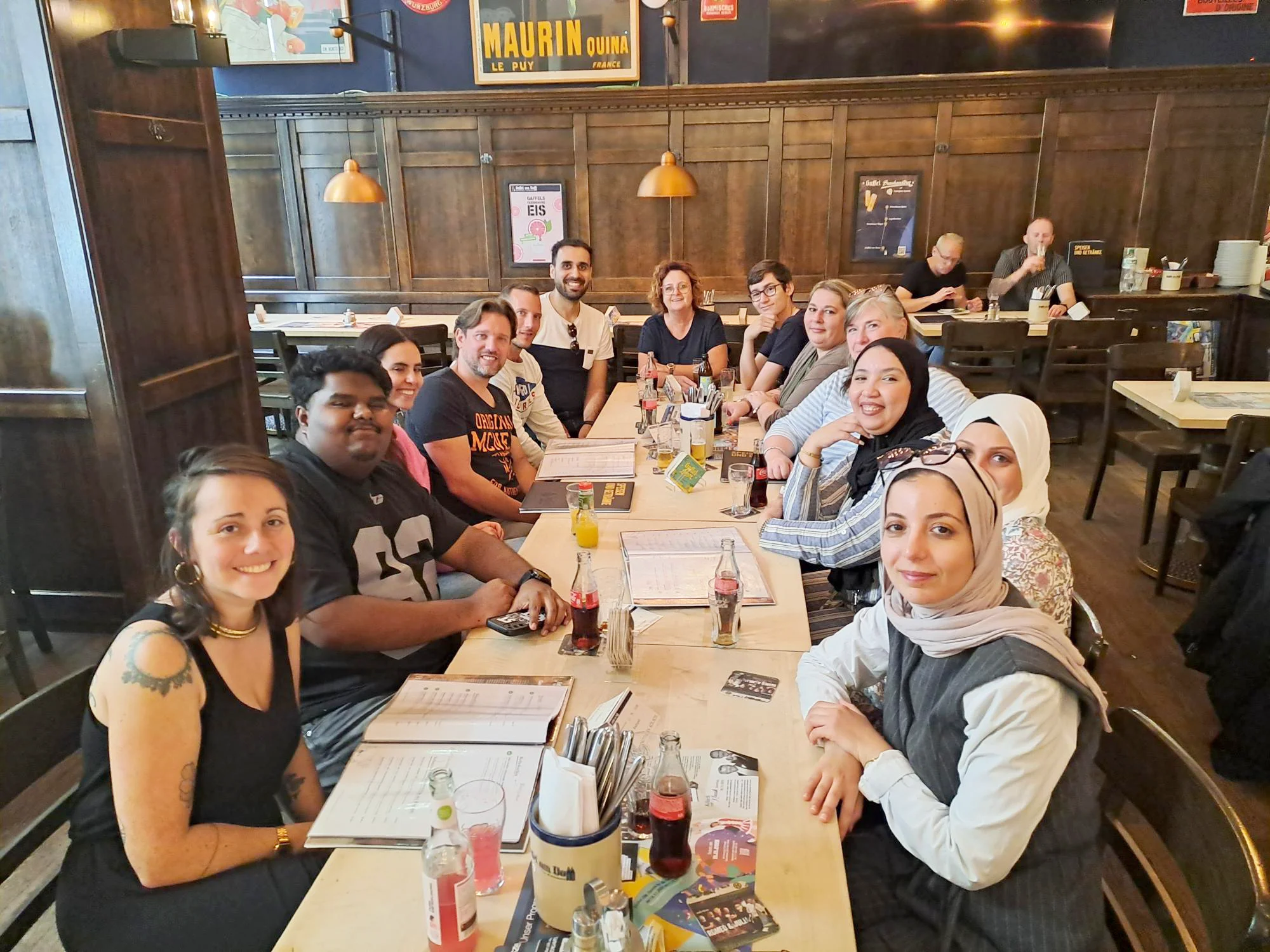 Auf dem Foto ist eine Gruppe von 13 Erwachsenen zu sehen, die an zwei zusammengeschobenen Tischen in einem Restaurant oder einer Gaststätte sitzen. Vor ihnen stehen Getränke und es liegen Speisekarten auf dem Tisch. Im Hintergrund sind weitere Tische, Holzwände und Bilder zu erkennen.