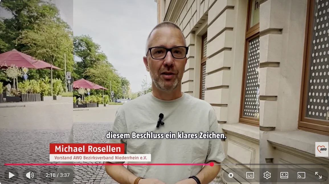 Auf dem Bild ist ein Mann zu sehen, der in einer städtischen Umgebung in die Kamera spricht und offenbar ein Statement abgibt. Bildbeschreibung Der Mann steht auf einem gepflasterten Platz oder einer Straße mit Bäumen und Außengastronomie (rote Sonnenschirme) im Hintergrund. Rechts im Bild ist die Fassade eines Gebäudes mit Fensterreihe zu sehen, die wie eine Verwaltungs- oder Bildungsinstitution wirkt. Er trägt ein helles T‑Shirt, eine Brille und ein am Kragen befestigtes Ansteck‑Mikrofon, was darauf hindeutet, dass es sich um ein professionell produziertes Interview oder Statement-Video handelt. Unten eingeblendet sind Bauchbinde und Untertitel, die seinen Namen und seine Funktion nennen sowie den gesprochenen Text zusammenfassen. Zudem sieht man die Video-Steuerleiste mit Laufzeit (2:18 / 3:37), Lautstärke-Icon und weiteren Player-Buttons, sowie ein Logo unten rechts, das auf den Absender des Videos hinweist.