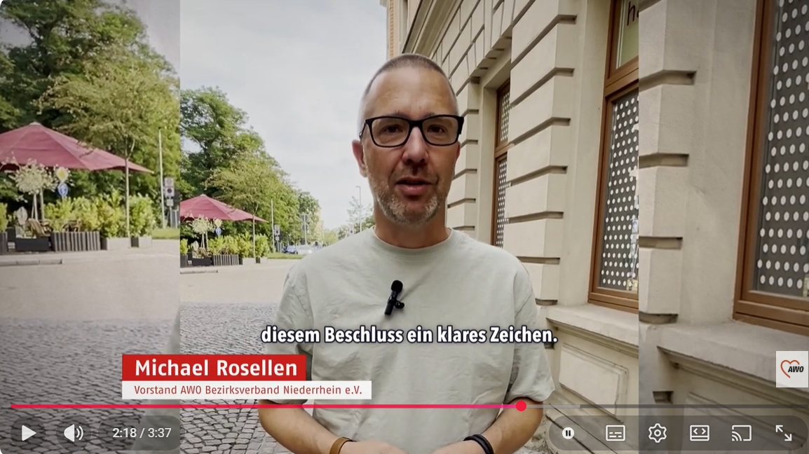 Auf dem Bild ist ein Mann zu sehen, der in einer städtischen Umgebung in die Kamera spricht und offenbar ein Statement abgibt. Bildbeschreibung Der Mann steht auf einem gepflasterten Platz oder einer Straße mit Bäumen und Außengastronomie (rote Sonnenschirme) im Hintergrund. Rechts im Bild ist die Fassade eines Gebäudes mit Fensterreihe zu sehen, die wie eine Verwaltungs- oder Bildungsinstitution wirkt. Er trägt ein helles T‑Shirt, eine Brille und ein am Kragen befestigtes Ansteck‑Mikrofon, was darauf hindeutet, dass es sich um ein professionell produziertes Interview oder Statement-Video handelt. Unten eingeblendet sind Bauchbinde und Untertitel, die seinen Namen und seine Funktion nennen sowie den gesprochenen Text zusammenfassen. Zudem sieht man die Video-Steuerleiste mit Laufzeit (2:18 / 3:37), Lautstärke-Icon und weiteren Player-Buttons, sowie ein Logo unten rechts, das auf den Absender des Videos hinweist.