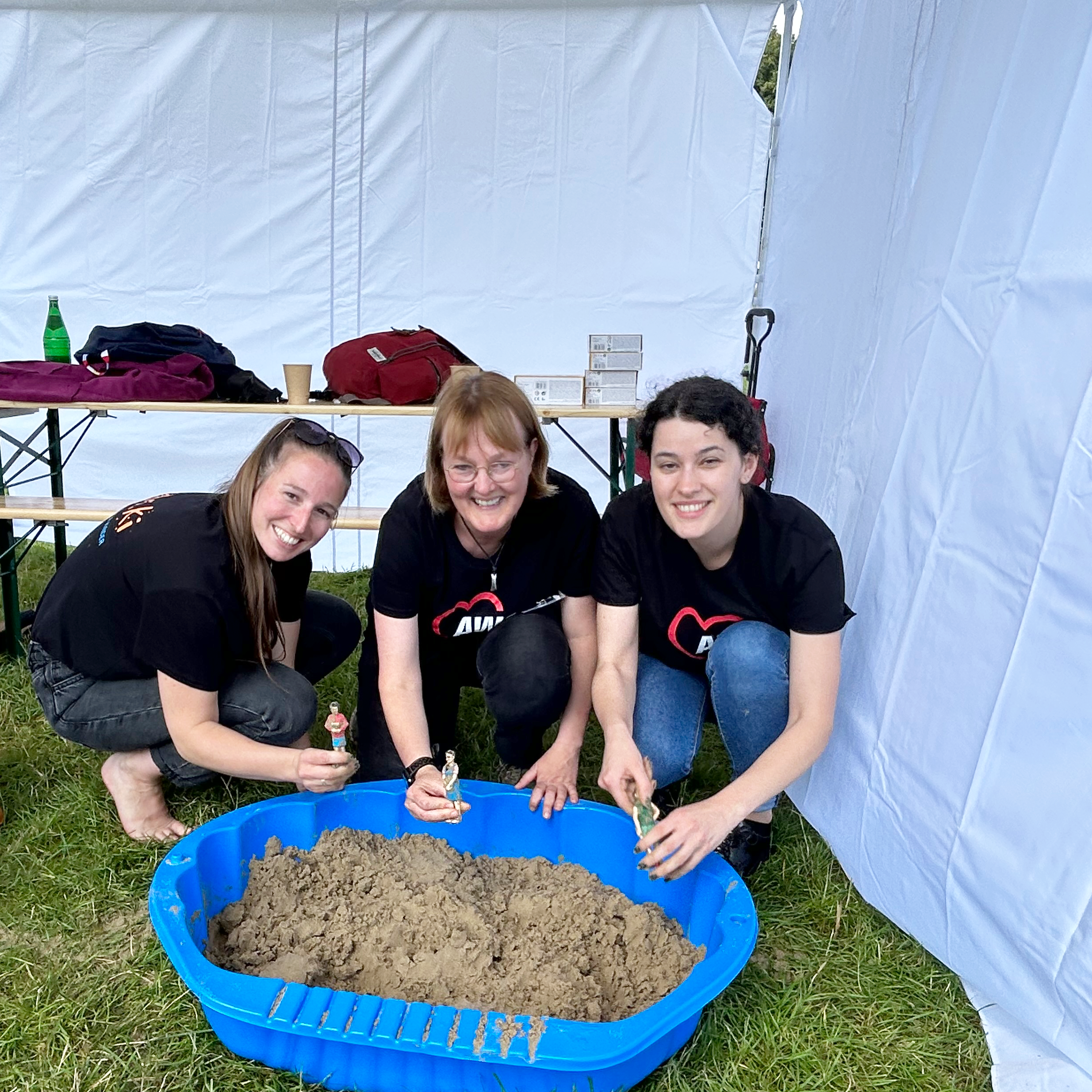 Auf dem Bild sind drei Personen zu sehen. Sie knien auf dem Gras vor einem weißen Zelt. Die Personen tragen schwarze T-Shirts mit einem roten Logo auf der Brust. Vor ihnen steht eine blaue Plastikwanne, die mit Sand gefüllt ist.