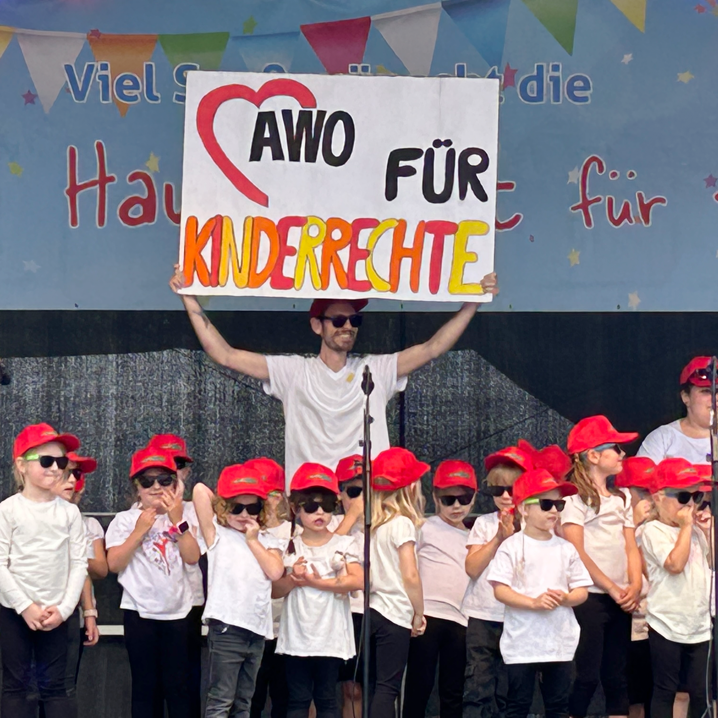Auf dem Bild ist eine Gruppe von Kindern. Sie stehen vor einer Bühne mit einem Banner. Eine Person hält ein großes Schild über dem Kopf, auf dem “AWO FÜR KINDERRECHTE” steht. 