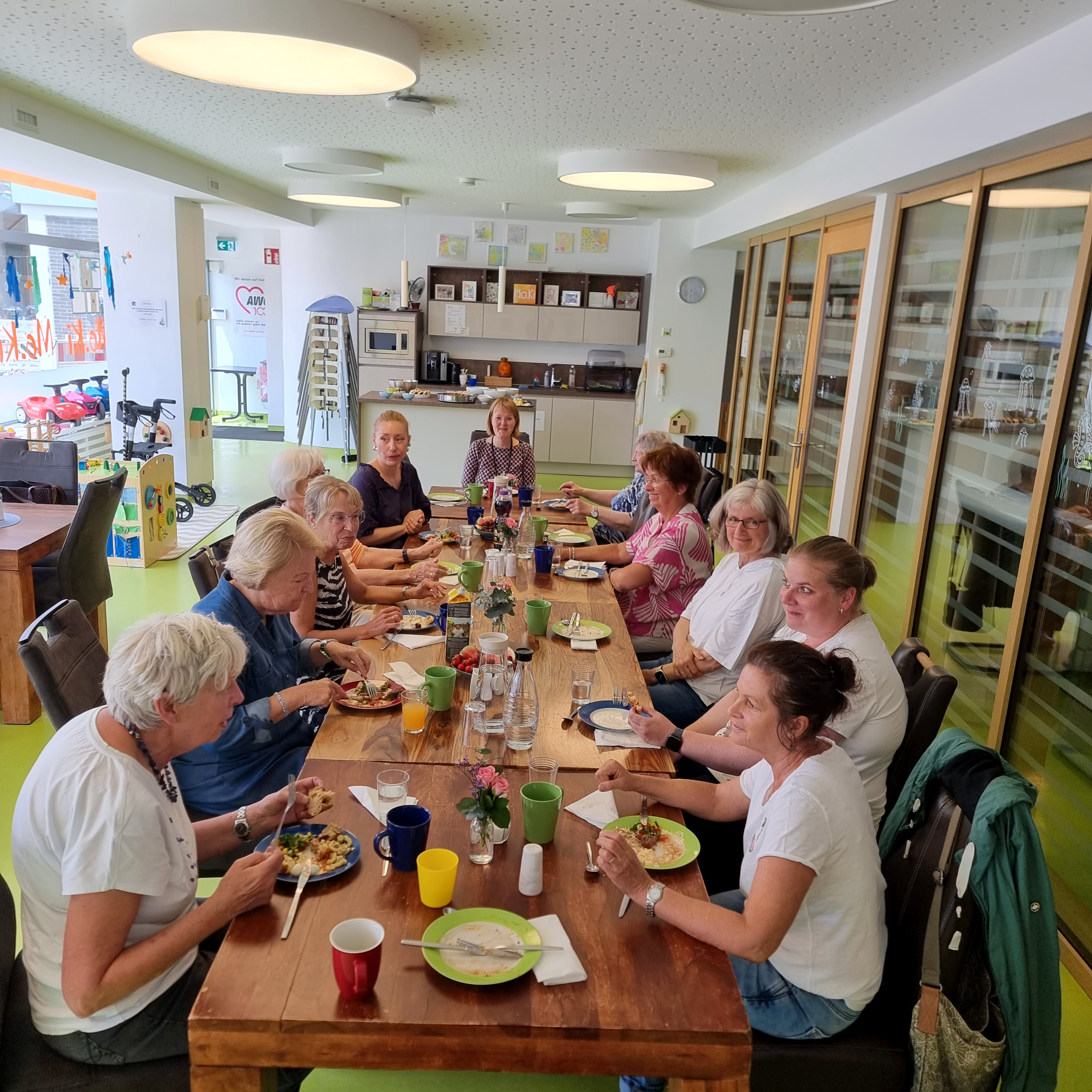 Auf dem Foto ist eine Gruppe von etwa 14 Frauen zu sehen, die gemeinsam an einem langen Holztisch in einem freundlich gestalteten Gemeinschaftsraum essen. Der Raum wirkt modern und einladend, mit großen Fenstern auf der rechten Seite und einer Küchenzeile im Hintergrund. Auf dem Tisch stehen bunte Tassen, Teller mit Essen, Gläser und Karaffen mit Wasser. Die Atmosphäre wirkt freundlich und gesellig. Im Hintergrund sind Spielgeräte und Hinweise auf eine AWO-Einrichtung sichtbar (z. B. das AWO-Logo auf einem Plakat an der Tür), was auf ein generationsübergreifendes oder soziales Angebot schließen lässt – möglicherweise ein gemeinsames Essen oder ein Treffpunkt für Ehrenamtliche, Seniorinnen oder Teilnehmerinnen eines Projekts.