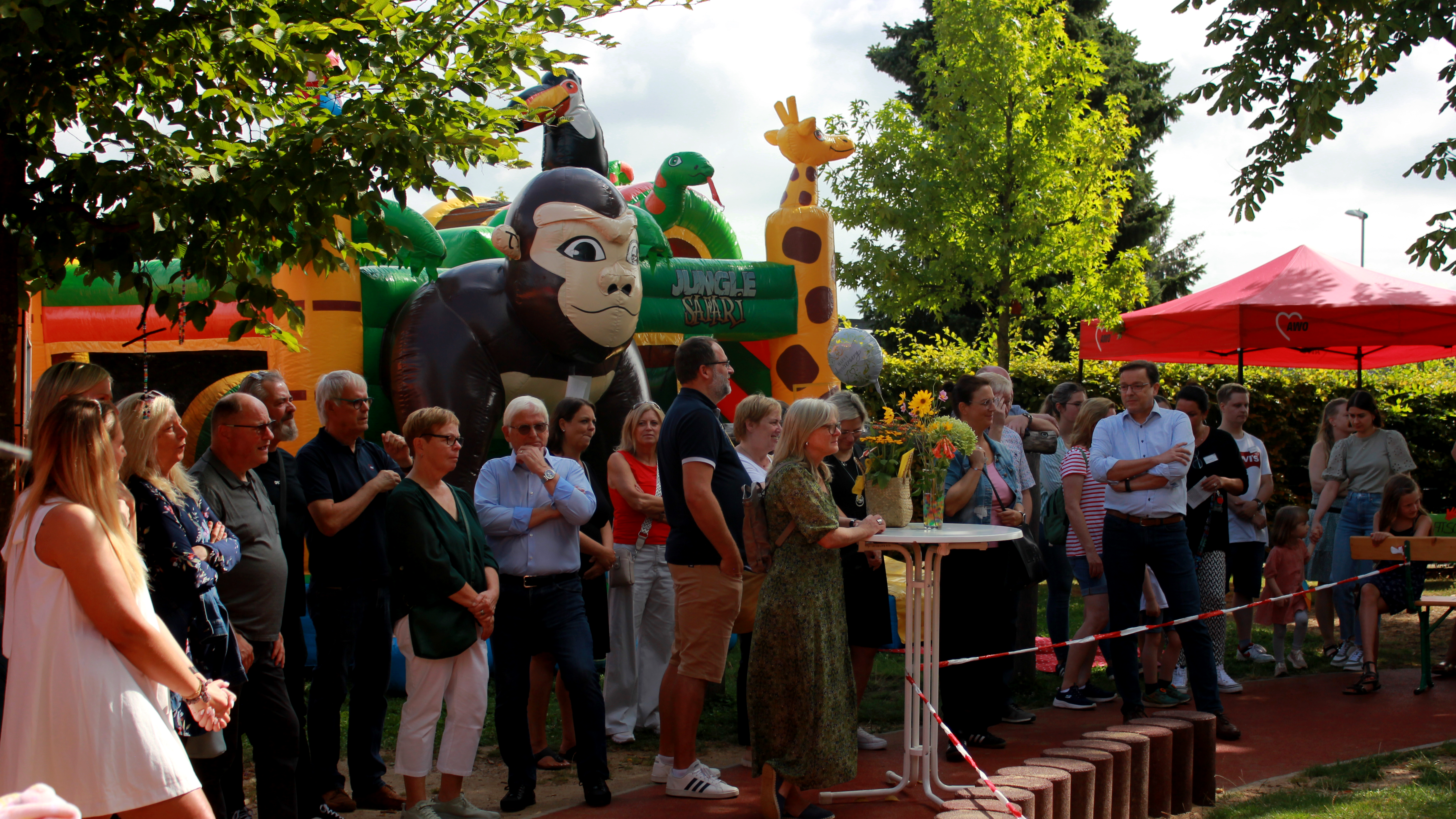 Auf dem Foto ist eine Outdoor-Veranstaltung zu sehen, bei der mehrere Personen versammelt sind. Im Hintergrund befindet sich eine große aufblasbare Struktur mit einem Gorilla und anderen bunten Elementen, die Teil eines Kinderspielbereichs sind. Im Vordergrund steht eine Gruppe von Personen, die etwas außerhalb des Bildes aufmerksam beobachtet. Die Umgebung wird von Bäumen und klarem Himmel geprägt.