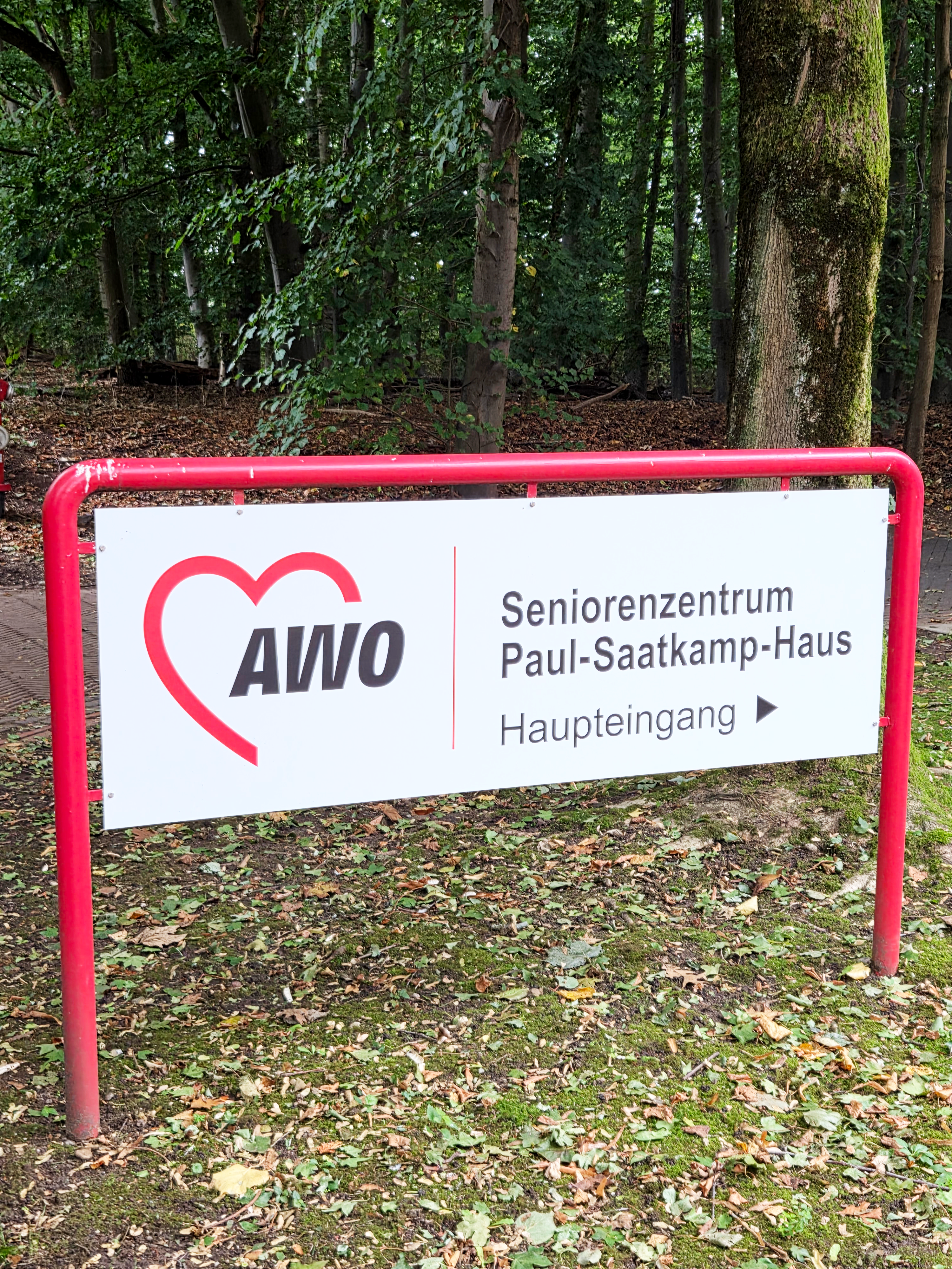  Auf dem Foto ist ein Schild zu sehen, das auf das "Seniorenzentrum Paul-Saatkamp-Haus" hinweist. Es hat einen weißen Hintergrund mit rotem Rahmen und roten Buchstaben. Auf der linken Seite des Schildes befindet sich das Logo der AWO (Arbeiterwohlfahrt), das aus einem stilisierten roten Herzen mit den Buchstaben "AWO" besteht. Auf der rechten Seite stehen die Worte "Seniorenzentrum Paul-Saatkamp-Haus" und darunter "Haupteingang" mit einem Pfeil, der nach rechts zeigt. Das Schild steht auf einem mit Blättern bedeckten Boden, und im Hintergrund sind Bäume zu erkennen, was darauf hindeutet, dass sich das Zentrum in einer grünen, waldähnlichen Umgebung befindet.