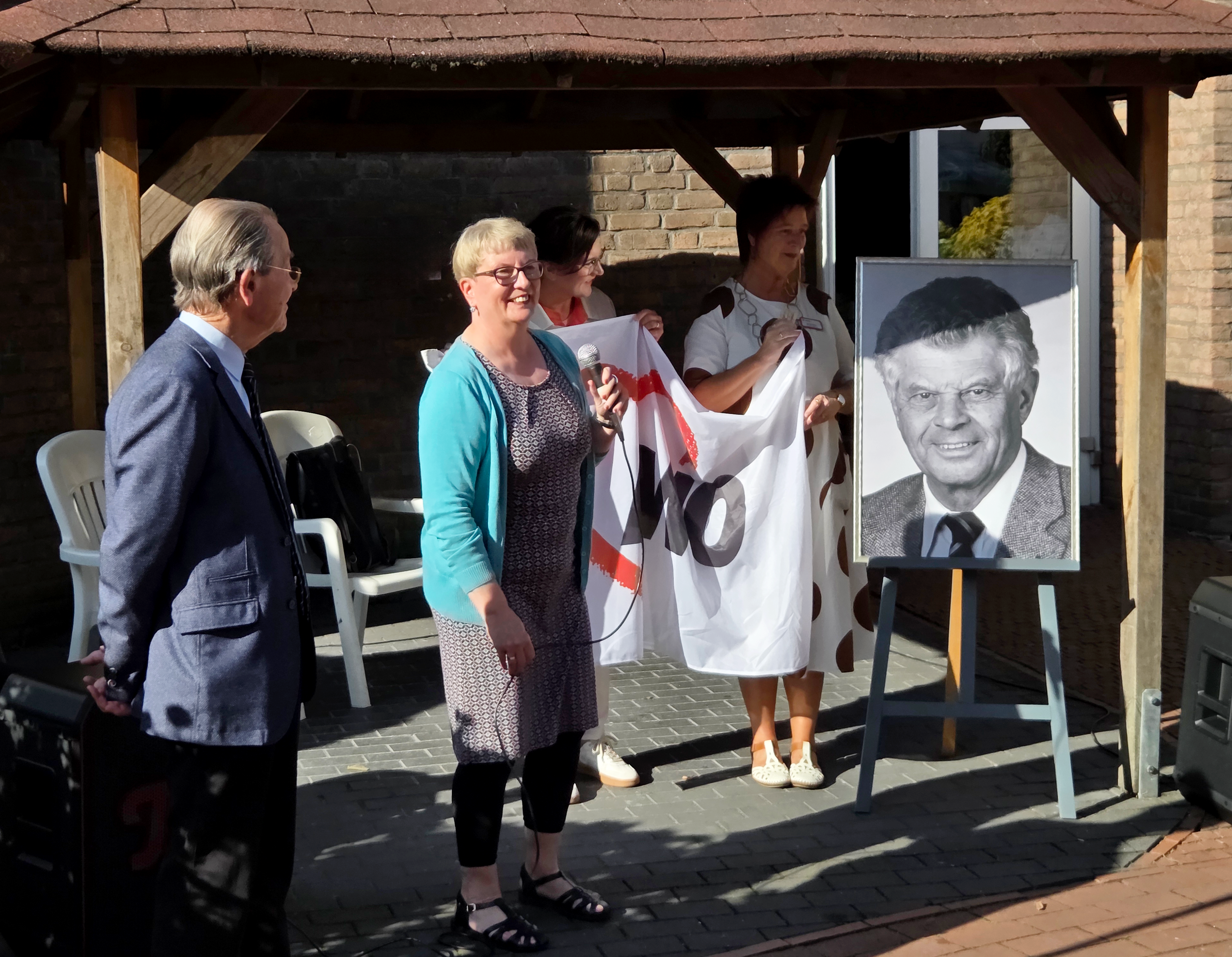 Auf dem Foto sind drei Personen zu sehen, die unter einem Unterstand stehen, vermutlich bei einer Zeremonie oder Veranstaltung im Freien. In der Mitte hält eine Frau, die eine blaue Strickjacke trägt, eine Art Fahne oder Banner, auf dem die Buchstaben "AWO" zu erkennen sind. Neben ihr, auf ihrer rechten Seite, steht eine andere Frau, die ein weißes, gepunktetes Kleid trägt und ebenfalls das Banner festhält. Eine dritte Person, ein Mann in einem blauen Anzug, steht auf der linken Seite. Neben den Personen steht eine Staffelei mit einem gerahmten Porträtfoto eines Mannes in einem Anzug. Die Szene spielt sich vor einer Ziegelsteinwand ab, und im Hintergrund sind weiße Plastikstühle zu sehen.