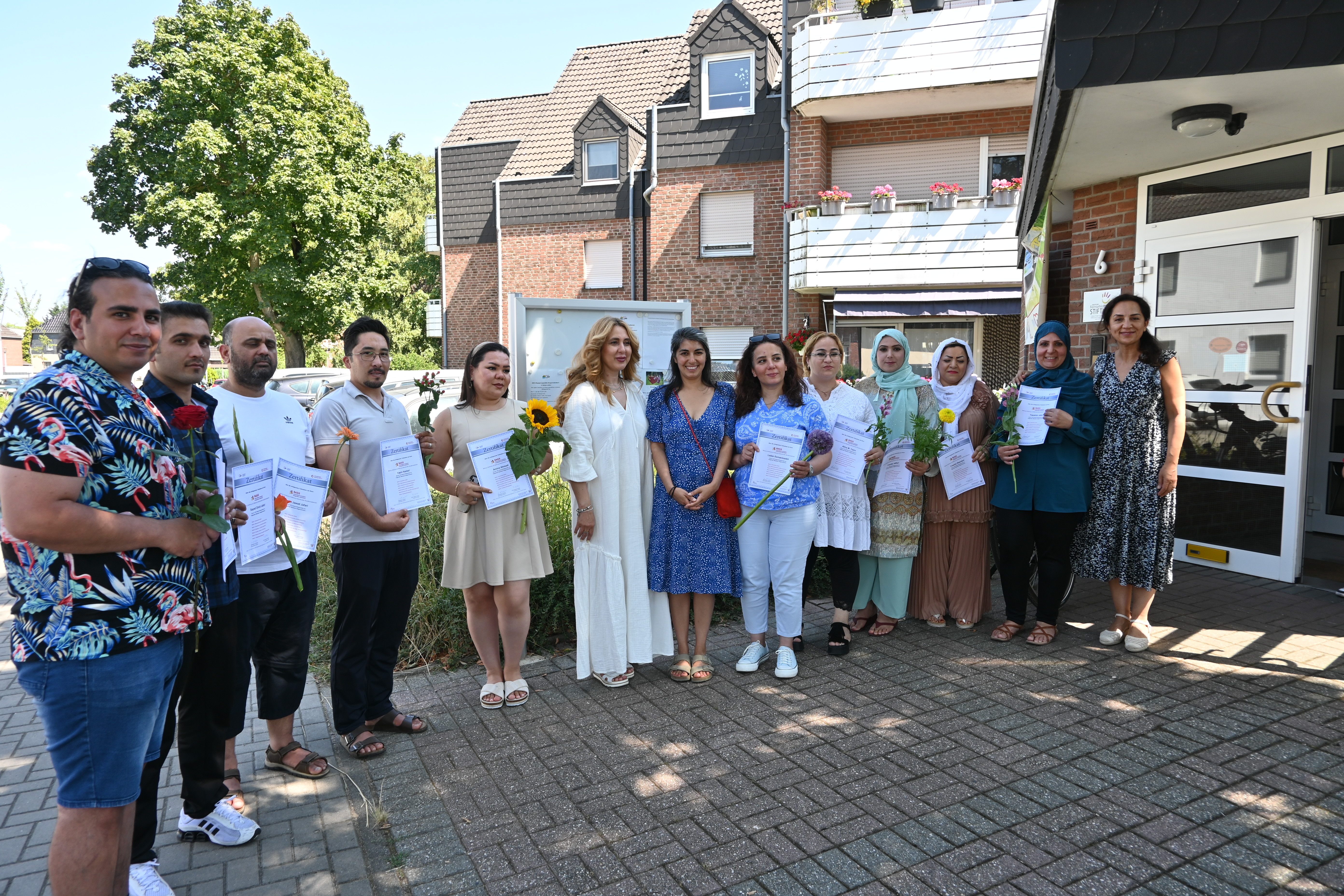Teilnehmende eines Bildungs- und Qualifizierungsprogramms präsentieren ihre Zertifikate und Blumen vor einem Wohn- und Gemeinschaftsgebäude. Das Gruppenfoto wurde im Freien bei sonnigem Wetter aufgenommen und dokumentiert den erfolgreichen Abschluss der Maßnahme.