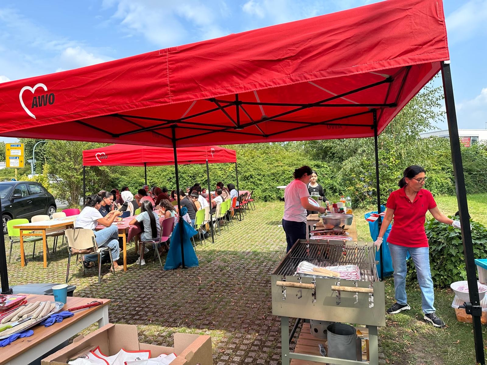 Das Bild zeigt eine fröhliche Outdoor-Veranstaltung unter mehreren roten Pavillons mit dem Logo der AWO (Arbeiterwohlfahrt). Menschen sitzen an Tischen, essen und unterhalten sich. Im Vordergrund steht ein Grill, an dem zwei Personen Speisen zubereiten. Die Atmosphäre wirkt entspannt und gemeinschaftlich – typisch für ein Sommerfest oder ein Nachbarschaftstreffen mit Essen, Gesprächen und Aktivitäten im Freien.