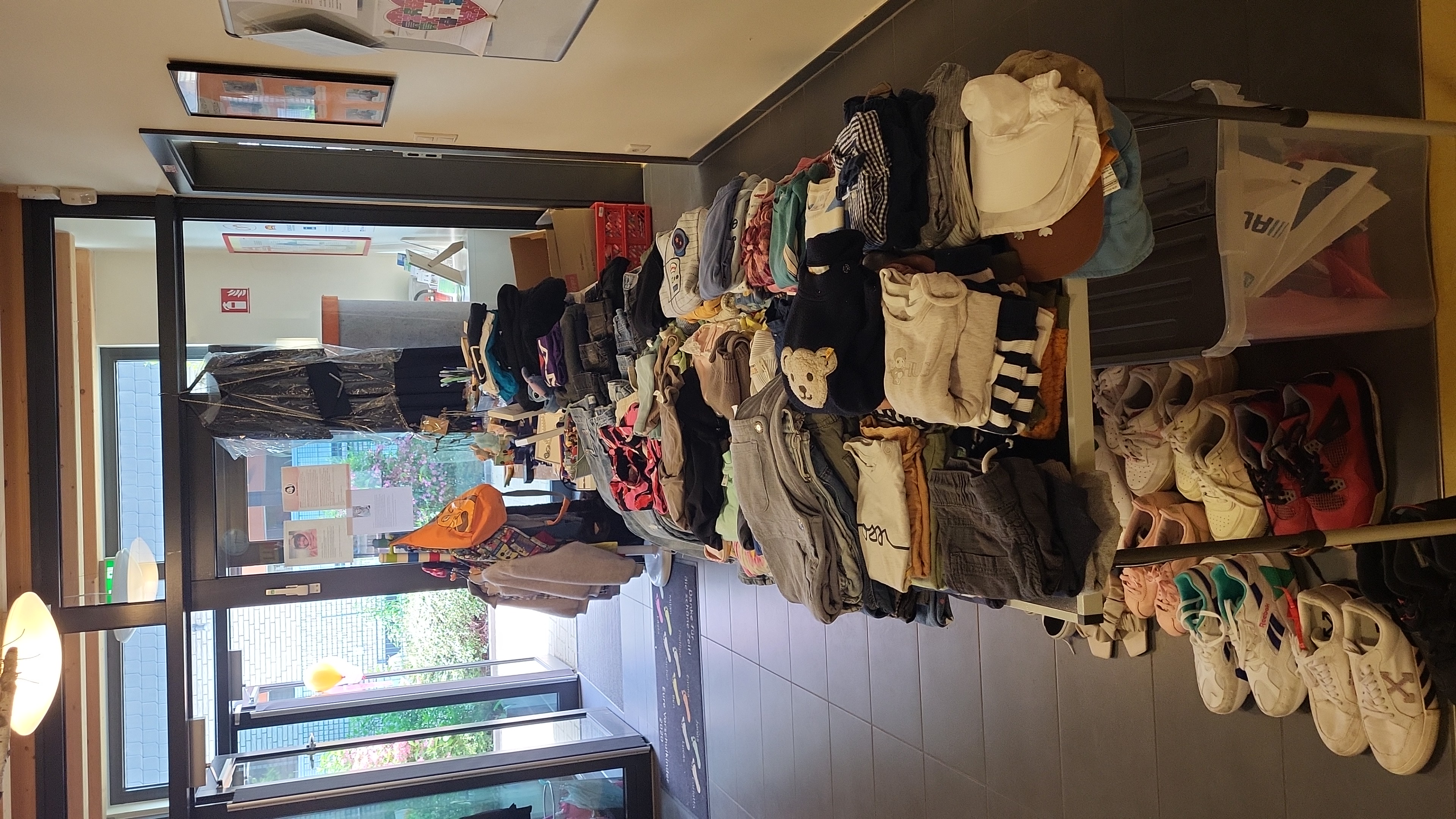 Auf dem Bild sieht man den Eingangs‑ oder Flurbereich eines Gebäudes, in dem auf mehreren Tischen und darunter Kleidung und Schuhe ausgelegt sind. Es wirkt wie eine kleine Kleiderbörse oder Spenden‑/Tauschaktion, vor allem mit gefalteten Kinder‑ und Erwachsenensachen, Schuhpaaren am Boden sowie einigen hängenden Kleidungsstücken und Infoplakaten an den Türen und Wänden.