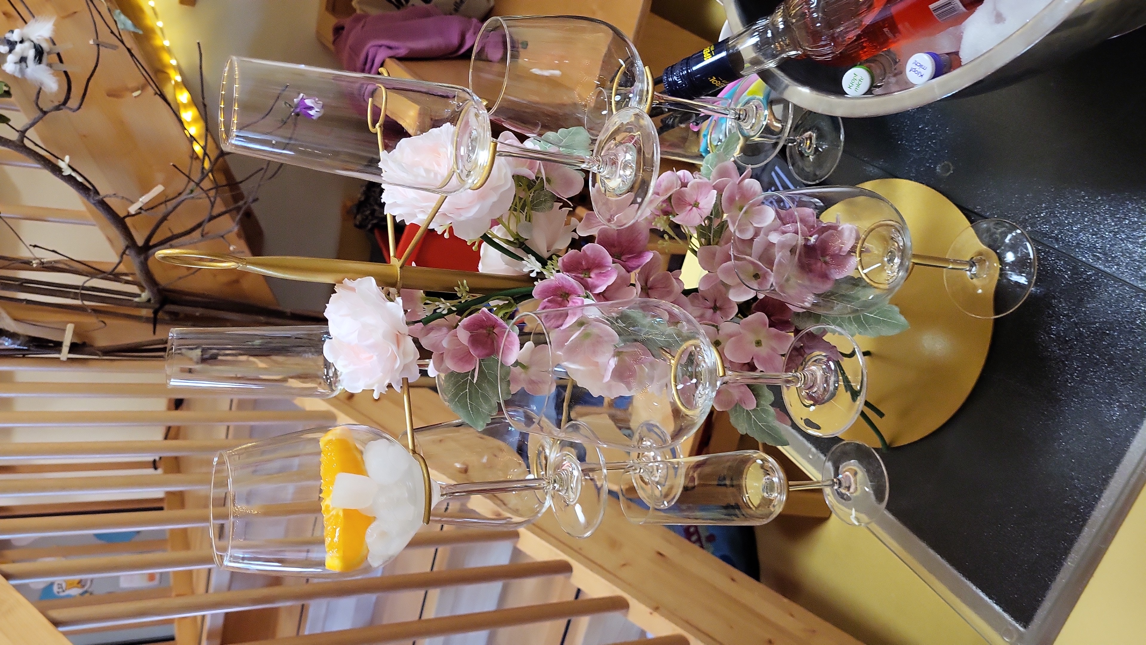 Auf dem Bild ist ein dekorativ arrangierter Glasständer zu sehen, auf dem mehrere leere Wein- und Sektgläser hängen bzw. stehen. In der Mitte sind künstliche rosa und weiße Blumen angebracht, daneben erkennt man einen Sekt-/Getränkekühler mit Flaschen, im Hintergrund ein Holztreppengeländer und etwas Deko (Zweige, Lichterkette).