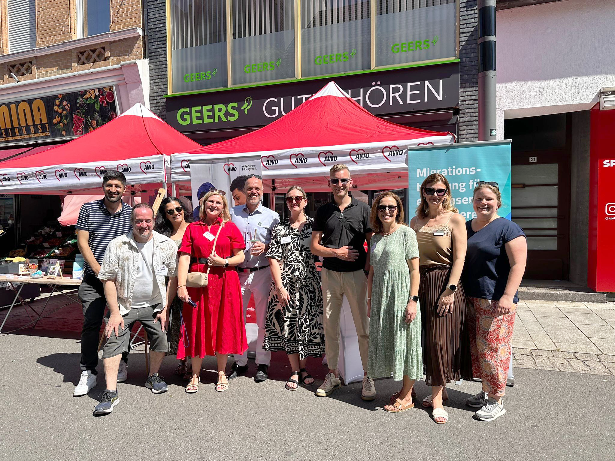 Auf dem Foto ist eine Gruppe von Menschen zu sehen, die gemeinsam vor einem Info-Stand der AWO (Arbeiterwohlfahrt) bei einer öffentlichen Veranstaltung stehen – vermutlich ein Straßen- oder Kulturfest. Der Stand ist mit roten Pavillons und AWO-Logos geschmückt. Ein Banner im Hintergrund weist auf das Thema „Migrationsberatung“ hin. Die Gruppe wirkt fröhlich und entspannt, viele lächeln in die Kamera. Es ist sonnig, die Menschen tragen sommerliche Kleidung. Im Hintergrund sind Geschäfte wie „GEERS GUT HÖREN“ und „MINA Feinkost“ zu erkennen. Das Bild vermittelt eine offene und gemeinschaftliche Atmosphäre. Wahrscheinlich handelt es sich um das „Fest der Kulturen“ in Grevenbroich, wie der Dateiname vermuten lässt.