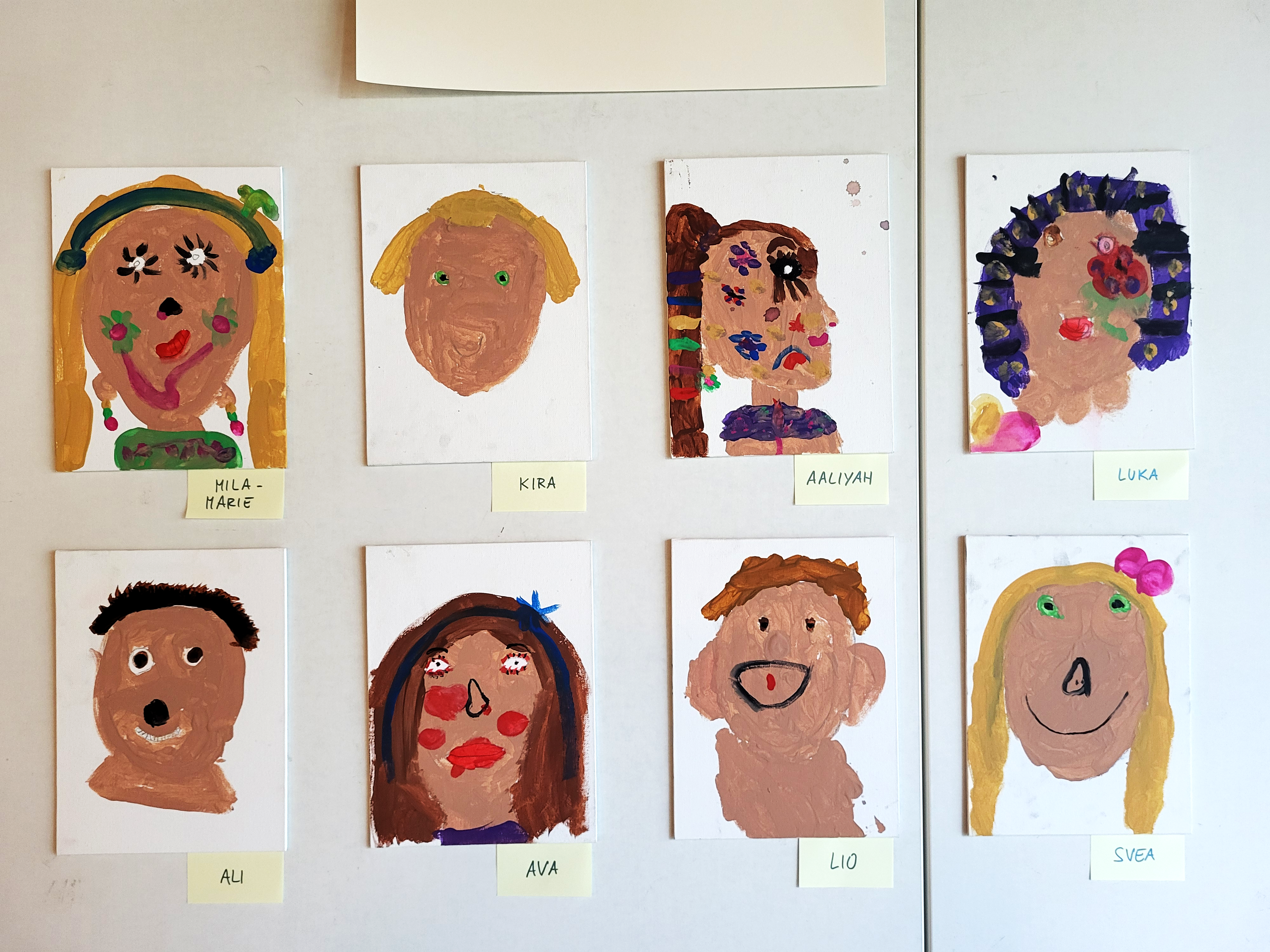 Auf dem Bild sind acht bunte Kinderporträts zu sehen, jeweils einzeln auf kleinen Leinwänden gemalt und an einer Wand aufgehängt. Unter jedem Bild klebt ein Zettel mit einem Vornamen (z.B. Mila‑Marie, Kira, Aaliyah, Luka, Ali, Ava, Lio, Svea), sodass erkennbar ist, welches Kind welches Bild gestaltet hat.