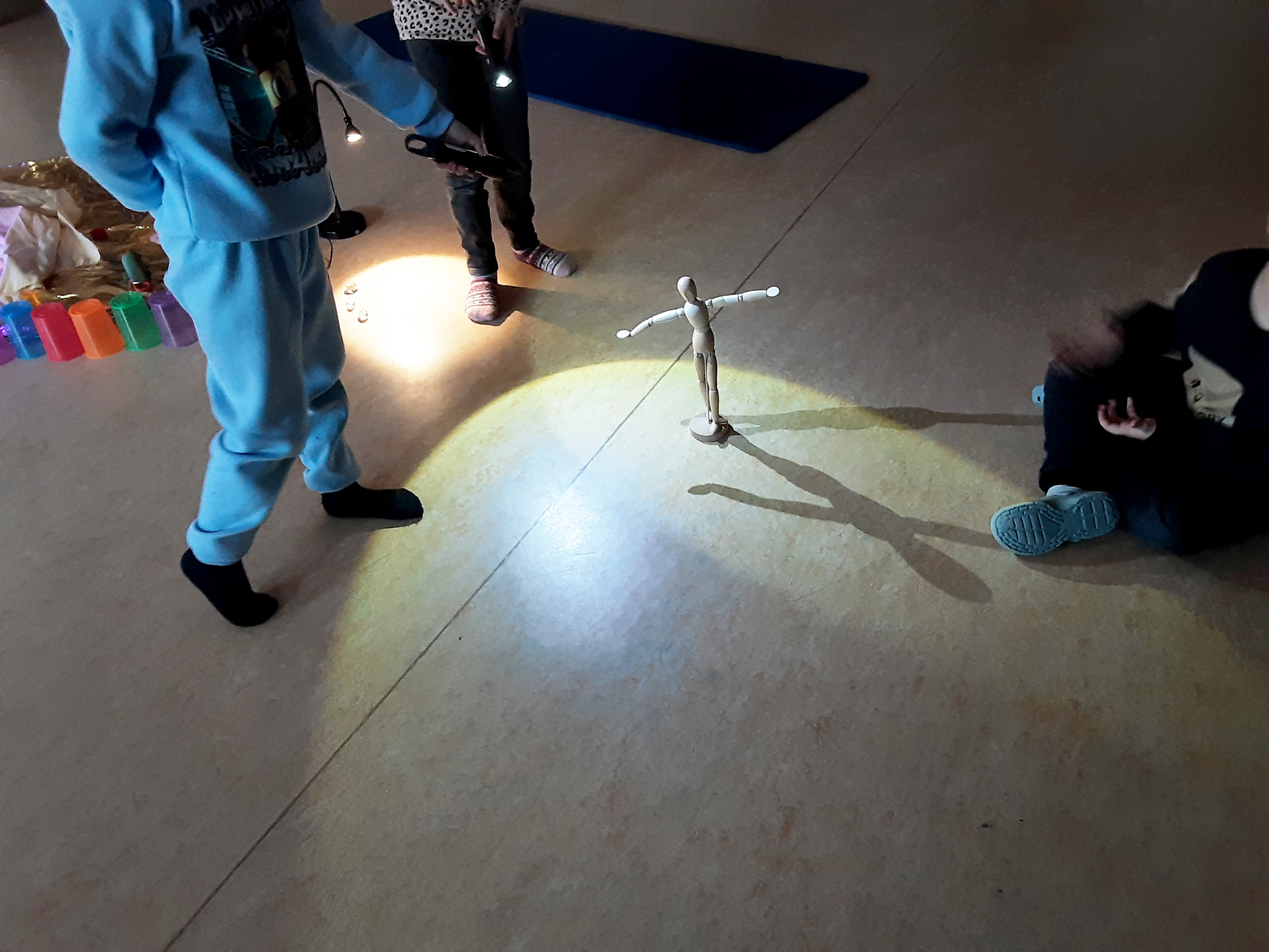 Auf dem Bild sind Kinder in einem Raum zu sehen, die mit Taschenlampen eine Holz‑Gliederpuppe anstrahlen. Die Puppe steht in der Mitte mit ausgebreiteten Armen und wirft durch das Licht deutlich sichtbare Schatten auf den Boden.