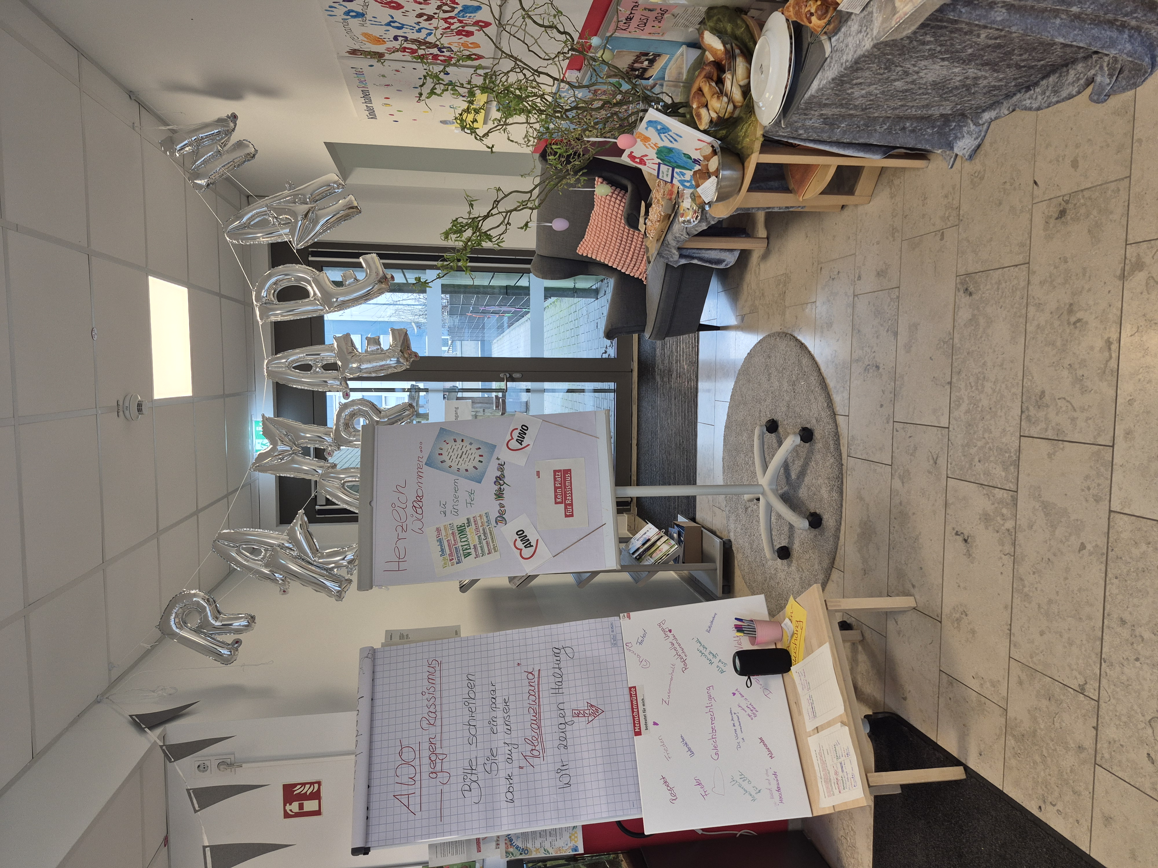 Das Bild zeigt den Eingangsbereich einer AWO-Kita, der anlässlich des Ramadan festlich dekoriert wurde. Im Einzelnen ist Folgendes zu sehen: Silberne Luftballon-Buchstaben an der Decke, die das Wort „RAMADAN" formen Zwei Flipcharts/Stellwände: Links: Ein Plakat mit der Aufschrift „AWO gegen Rassismus" – Besucher werden eingeladen, ein paar Worte auf die „Toleranzwand" zu schreiben, mit dem Satz „Wir zeigen Haltung" Rechts: Ein Willkommensschild mit „Herzlich Willkommen zu unserem Fest", mit AWO-Logos und dem Hinweis „Kein Platz für Rassismus" Eine beschriftete weiße Tafel auf dem Boden mit Begriffen wie Respekt, Frieden, Zusammenhalt, Gleichberechtigung usw. – offenbar eine interaktive Mitmach-Station zum Thema Menschenwürde Dekoration und Buffet auf der rechten Seite mit Gebäck, Tellern und einem Frühlingsast Kinderzeichnungen und Handabdrücke an der Wand rechts Ein gemütlicher Sessel mit bunten Kissen im Hintergrund