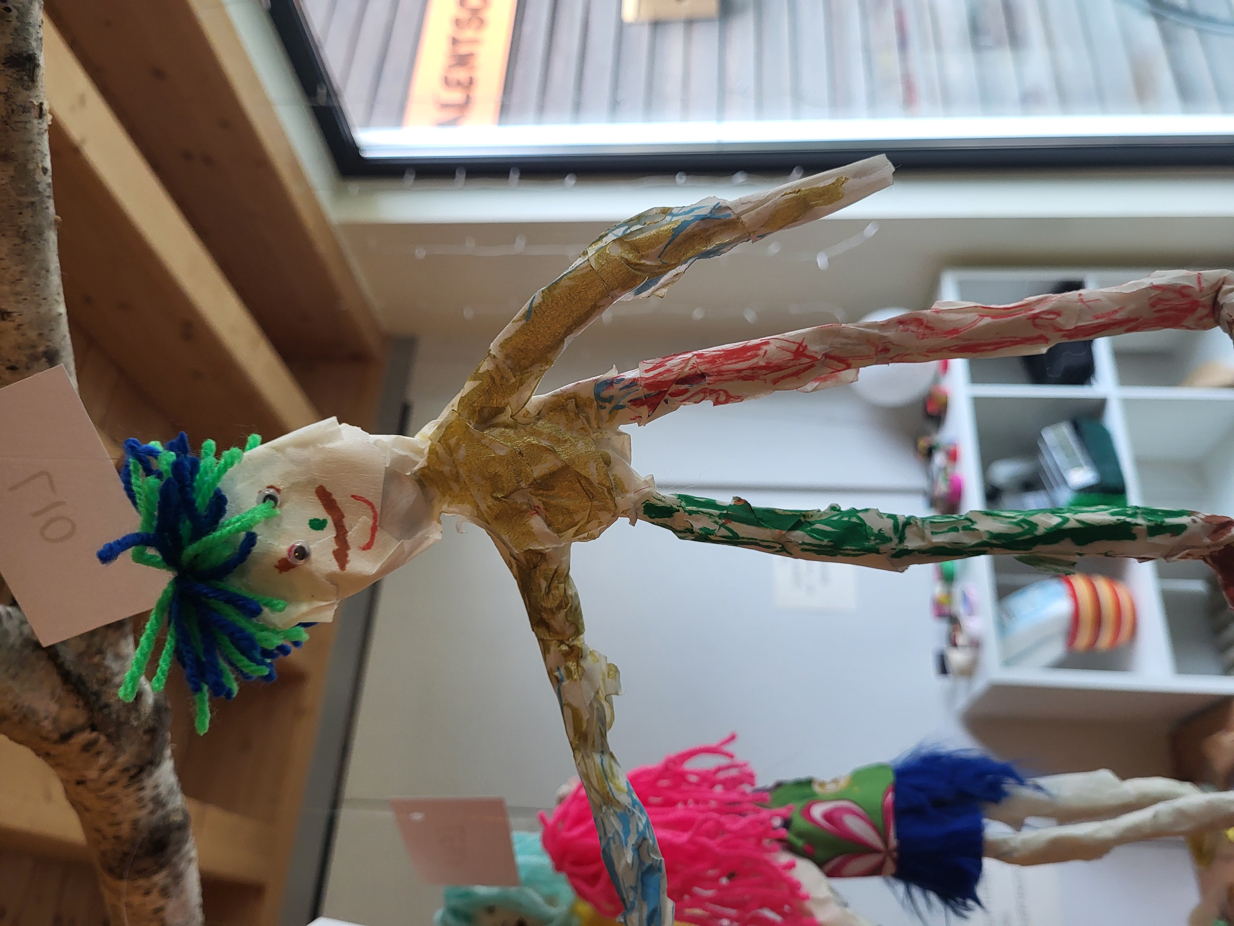 Auf dem Bild ist eine bunte, von Kindern gebastelte Figur aus Papier/Tape und Draht oder ähnlichem Material zu sehen, die an einem Ast von der Decke hängt. Die Figur hat Haare aus grüner und blauer Wolle, ein gemaltes Gesicht mit Mund und Nase sowie farbig bemalte Arme und Beine; oben hängt ein Schild mit dem Namen „Lio“.