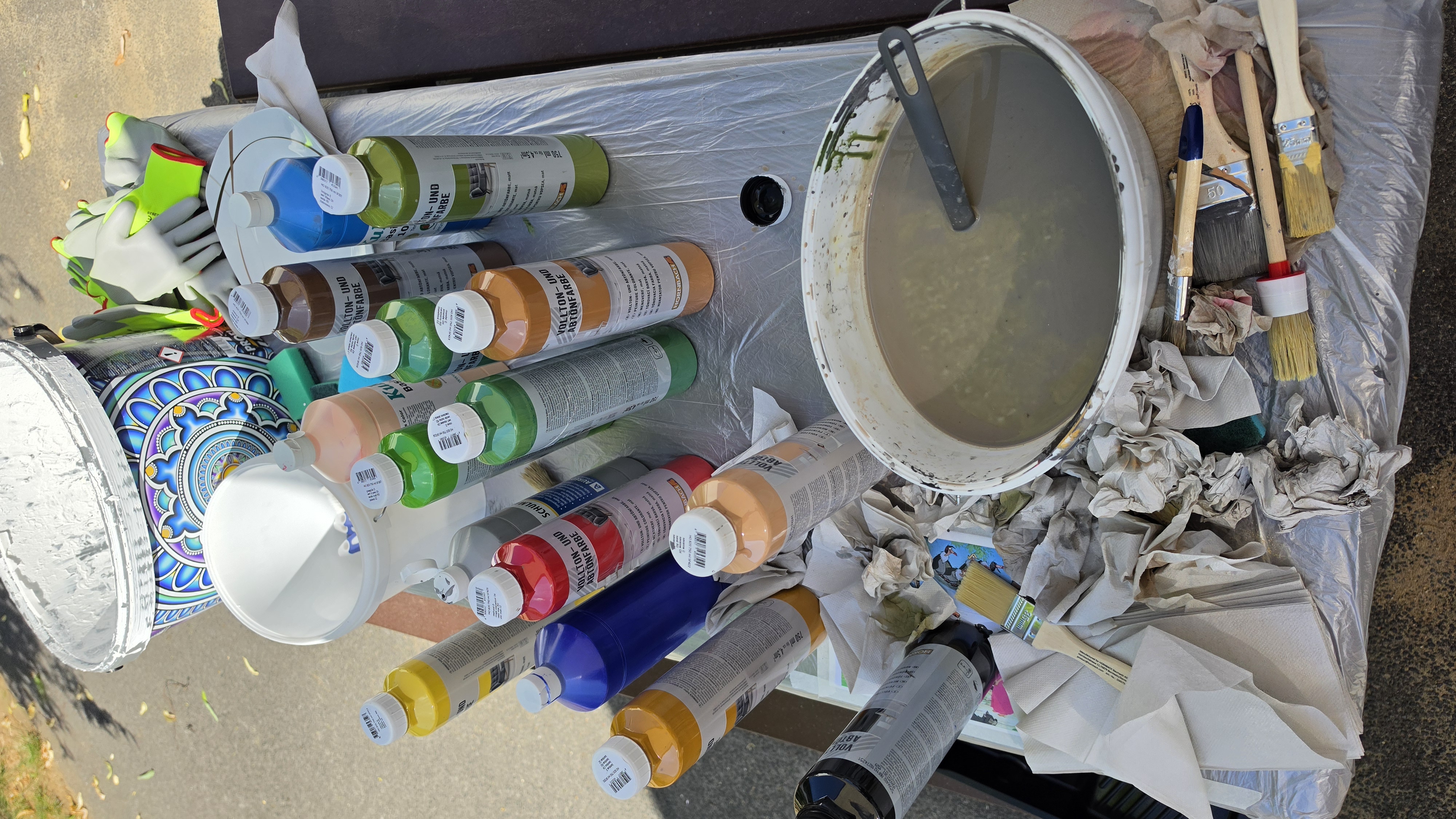 Auf dem Bild ist ein Arbeitstisch im Freien zu sehen, der für Mal‑ und Streicharbeiten vorbereitet ist. Auf dem Tisch stehen viele Flaschen mit bunten Acryl‑ oder Abtönfarben in verschiedenen Farben (Gelb, Blau, Rot, Grün, Braun usw.), daneben Eimer und Schalen mit Wasser bzw. verdünnter Farbe. Vorne liegen Pinsel und zusammengeknüllte Papiertücher, im Hintergrund erkennt man einen großen Farbkübel mit dekorativem Muster sowie Arbeitshandschuhe – insgesamt also ein kompletter Maler‑/Graffiti‑Arbeitsplatz für ein kreatives Projekt.