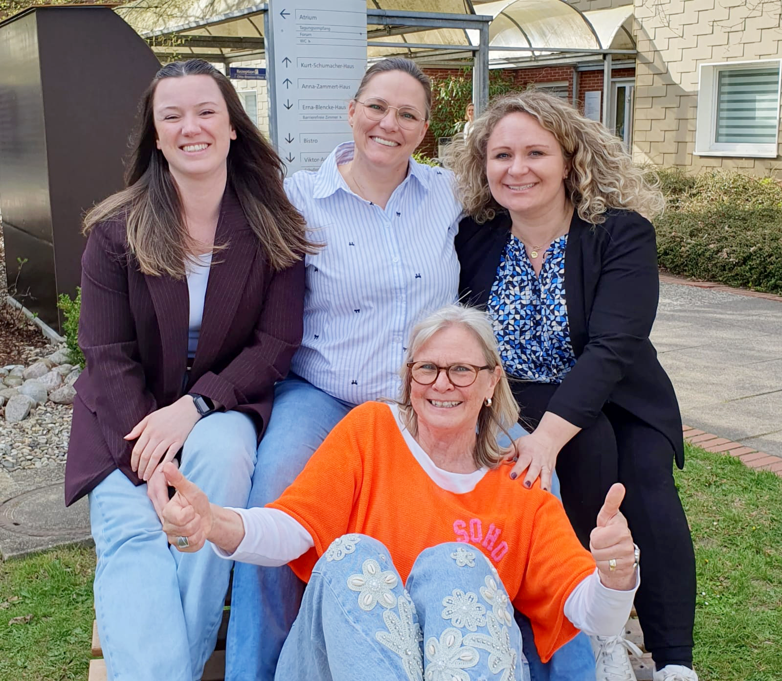  Was ist auf dem Bild zu sehen? Anhang 2026-Abschluss-der-Kita-Management-Fortbildung.jpg Auf dem Bild sitzen vier Personen draußen auf einer Bank oder einem niedrigen Sitzmöbel in einem Garten- bzw. Hofbereich. Drei Personen sitzen hinten nebeneinander, die vordere Person sitzt quer davor. Die vordere Person trägt ein auffällig orangenes Oberteil und zeigt mit beiden Händen den Daumen nach oben. Die Personen hinten tragen eher sportlich‑schicke Alltagskleidung (z.B. Blazer, Bluse, Hemd, Jeans). Im Hintergrund sind ein Weg, Grünflächen, ein Gebäude und ein großes, senkrechtes Hinweisschild mit mehreren Wegweisern zu sehen.