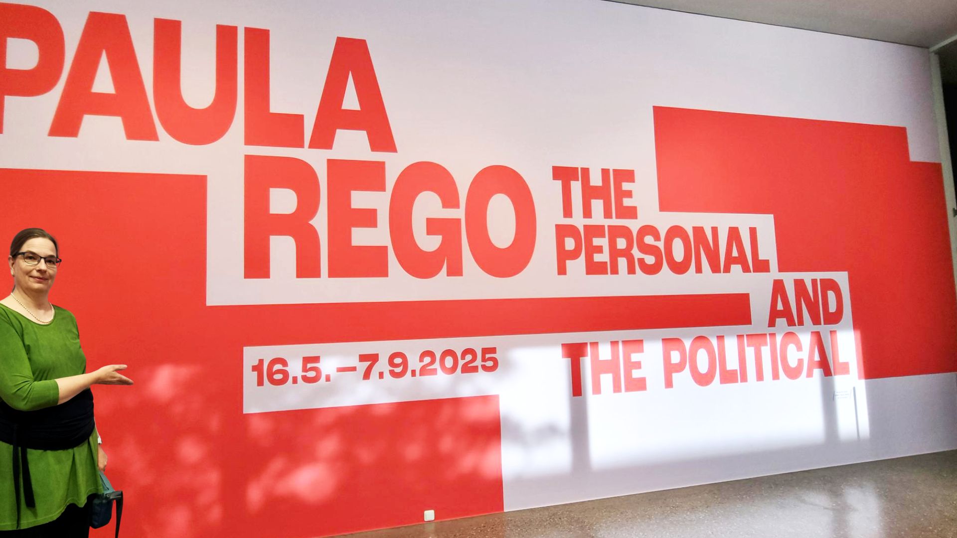 Auf dem Foto ist eine Frau zu sehen, die vor einer großen roten und weißen Wandinstallation steht. Auf der Wand ist in großen roten Buchstaben "PAULA REGO" zu lesen, gefolgt von kleinerem Text, der die Ausstellung als "THE PERSONAL AND THE POLITICAL" beschreibt. Auch das Datum der Ausstellung, "16.5.–7.9.2025", ist angegeben.