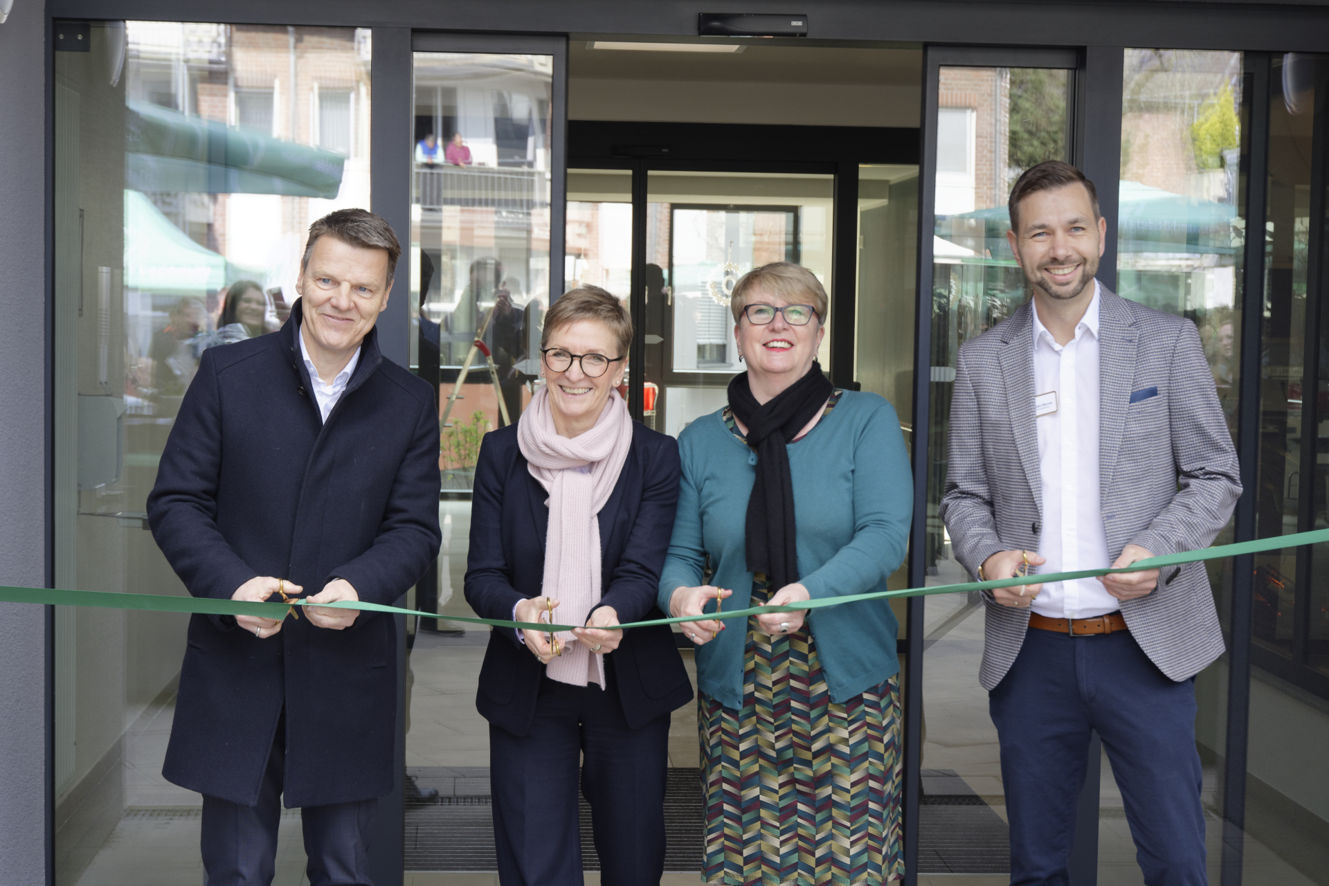 Auf dem Foto sieht es so aus, als ob eine feierliche Eröffnung stattfindet. Vier Personen stehen vor einem Gebäude und schneiden gemeinsam ein grünes Band durch – eine klassische Szene für eine Einweihungszeremonie. Alle scheinen gut gelaunt zu sein und lächeln in die Kamera.