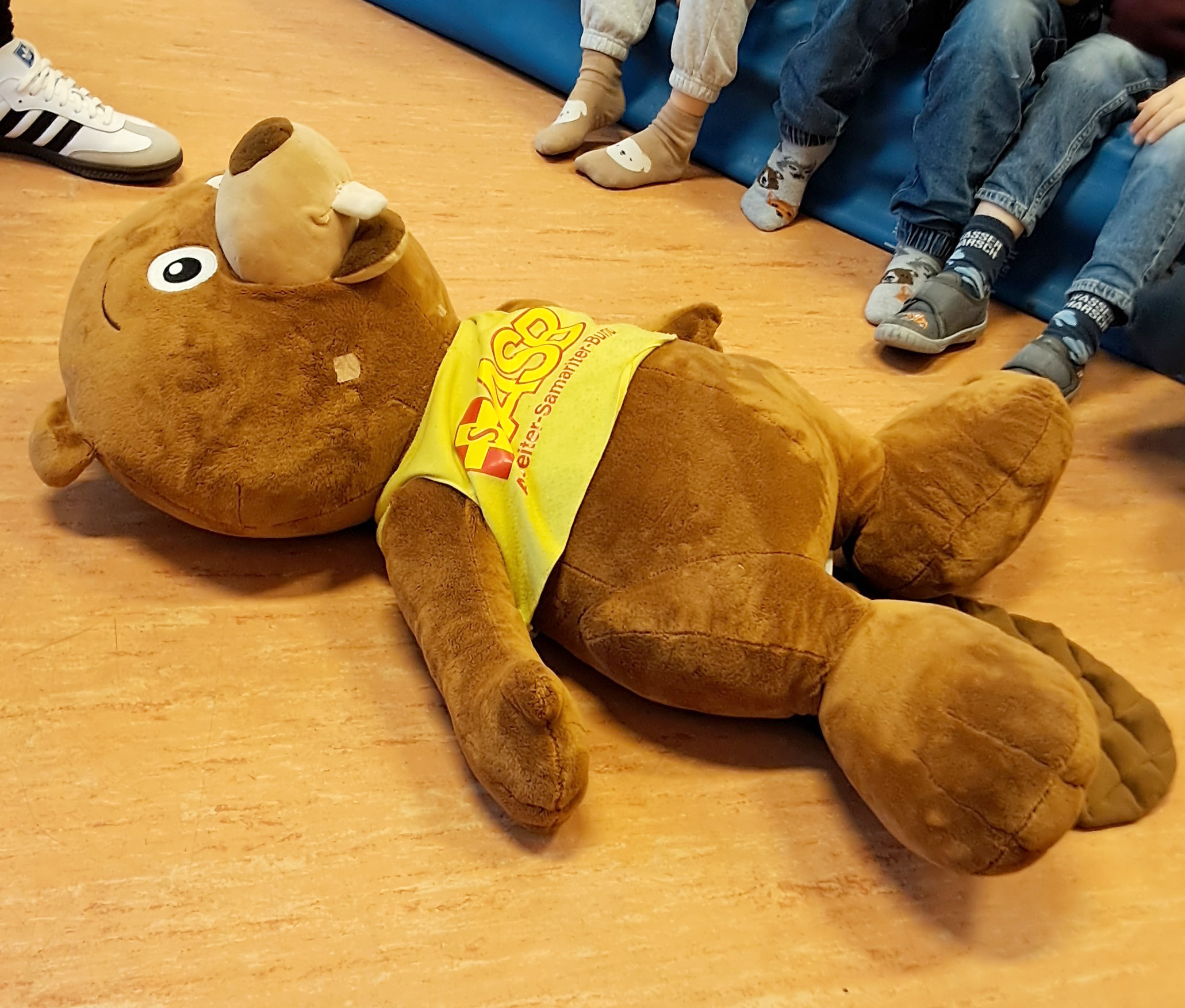 Auf dem Bild ist ein großes Plüschtier in Form eines Teddys zu sehen. Hier die Details: Ein braunes Teddy-Plüschtier mit charakteristischen großen Augen, einem freundlichen Gesichtsausdruck und einem auffälligen Geweih liegt auf dem hellen Holzboden. Das Stofftier trägt eine gelbe Schärpe/Binde mit der Aufschrift "ASB" und dem Text "Helfen Samariterbund" - dies ist das Maskottchen des Arbeiter-Samariter-Bundes. Im Hintergrund sieht man mehrere Kinder (ihre Beine und Füße in Jeans und verschiedenen Socken), die auf einer blauen Turnmatte sitzen. Links oben im Bild sind weiße Sneakers (wahrscheinlich die des Erwachsenen vom vorherigen Bild) zu erkennen. Die Szene spielt weiterhin in demselben Raum mit hellem Holzboden
