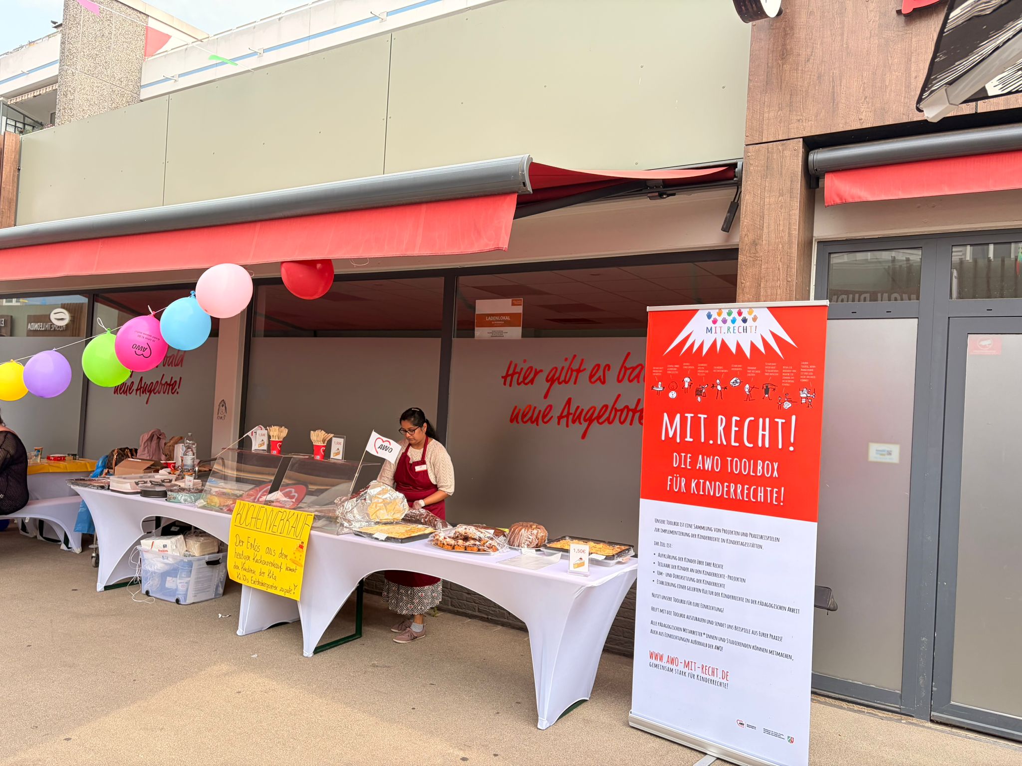 Auf dem Foto ist ein Aktionsstand der AWO vor einem Gebäude zu sehen. Links stehen mehrere mit weißen Tischhussen verkleidete Tische mit Kuchen, Gebäck und weiteren Speisen sowie einem handgeschriebenen Schild „Kuchenverkauf“, darüber sind bunte Luftballons aufgehängt. Rechts im Bild steht ein großer rot-weißer Aufsteller mit der Aufschrift „MIT.RECHT! Die AWO Toolbox für Kinderrechte!“. 