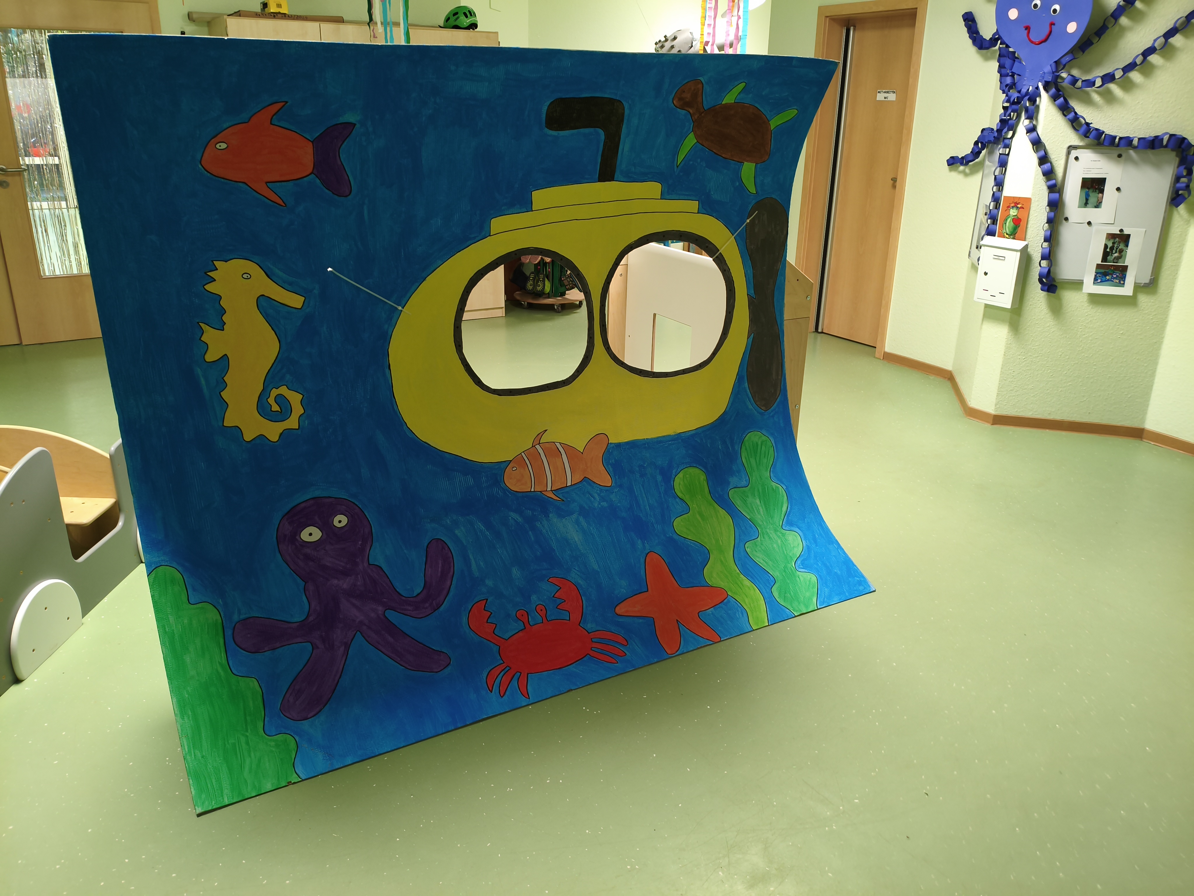Auf dem Bild steht in einem Kita‑Raum eine große bemalte Pappkulisse mit einem gelben U‑Boot in einer Unterwasserwelt. Darauf sind bunte Meerestiere wie Fisch, Seepferdchen, Krake, Krebs, Seestern, Schildkröte und Pflanzen zu sehen, und das U‑Boot hat zwei ausgeschnittene „Fenster“, durch die man hindurchschauen kann.