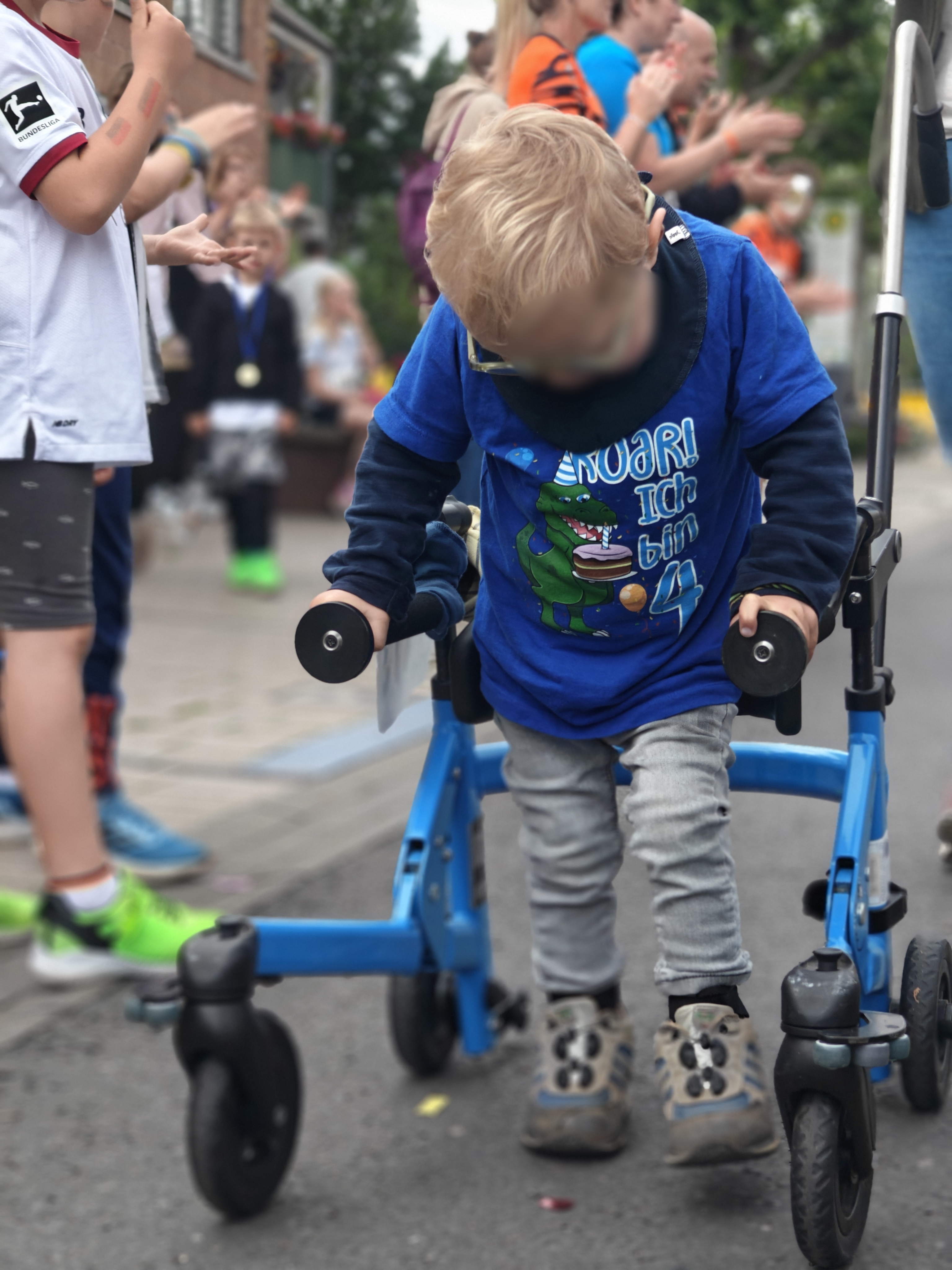 Im Mittelpunkt des Bildes ist ein kleiner Junge zu sehen, der mit einem blauen Gehtrainer auf einer abgesperrten Straße läuft. Er trägt ein blaues T‑Shirt mit einem Dinosaurier und dem Aufdruck „Roar! Ich bin 4“, dazu eine helle Hose und feste Schuhe. Links und im Hintergrund stehen weitere Kinder und Erwachsene am Straßenrand, die offenbar klatschen und anfeuern, was auf eine inklusive Lauf‑ oder Bewegungsaktion hindeutet.