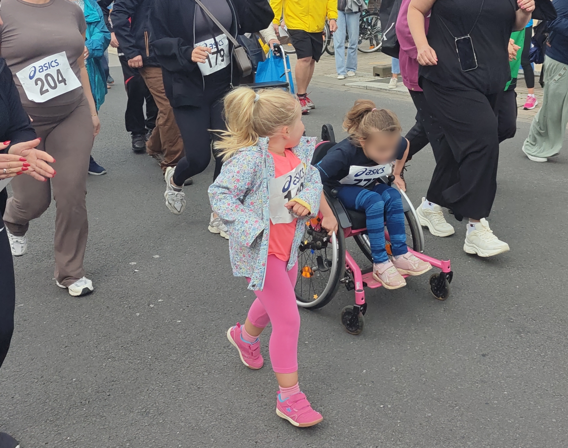 Auf dem Bild ist eine inklusive Laufszene bei einer Laufveranstaltung zu sehen. Im Mittelpunkt schiebt ein Mädchen im pinken Sportoutfit ein anderes Mädchen im Rollstuhl, beide tragen Startnummern eines Laufes. Rundherum laufen weitere Erwachsene und Kinder mit Startnummern auf der abgesperrten Straße mit, was den Charakter eines gemeinsamen Familien- oder Sponsorenlaufs unterstreicht.