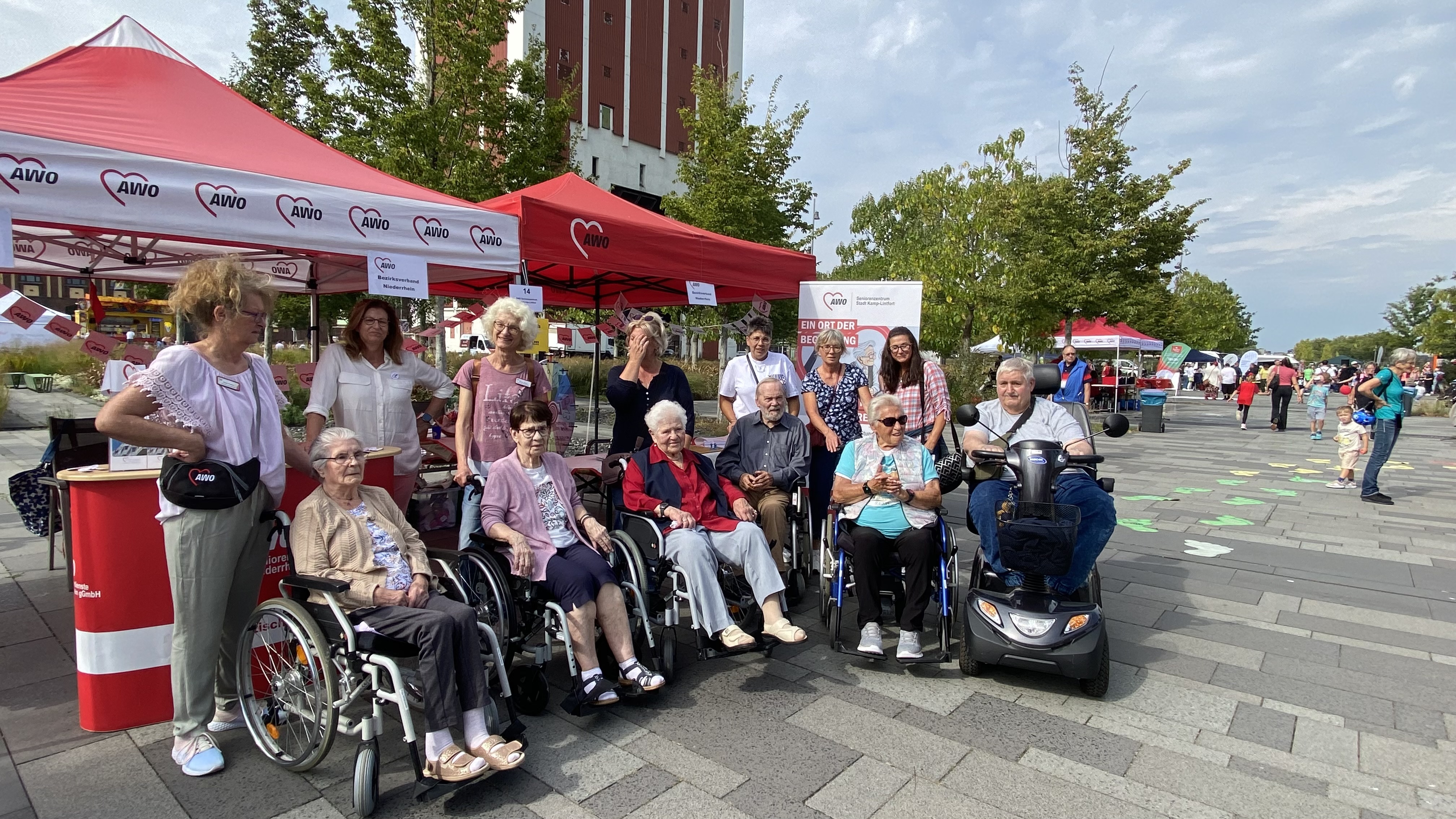  Auf dem Foto ist eine Gruppe von Personen zu sehen, die an einem AWO-Stand im Freien stehen und sitzen. Mehrere der Personen sitzen in Rollstühlen oder einem Elektromobil. Im Hintergrund sind zwei rote AWO-Pavillons, ein größeres Gebäude und weitere Menschen zu sehen, die sich auf einem Platz bewegen. Der Boden ist gepflastert und der Himmel ist bewölkt. Es scheint sich um eine öffentliche Veranstaltung zu handeln.