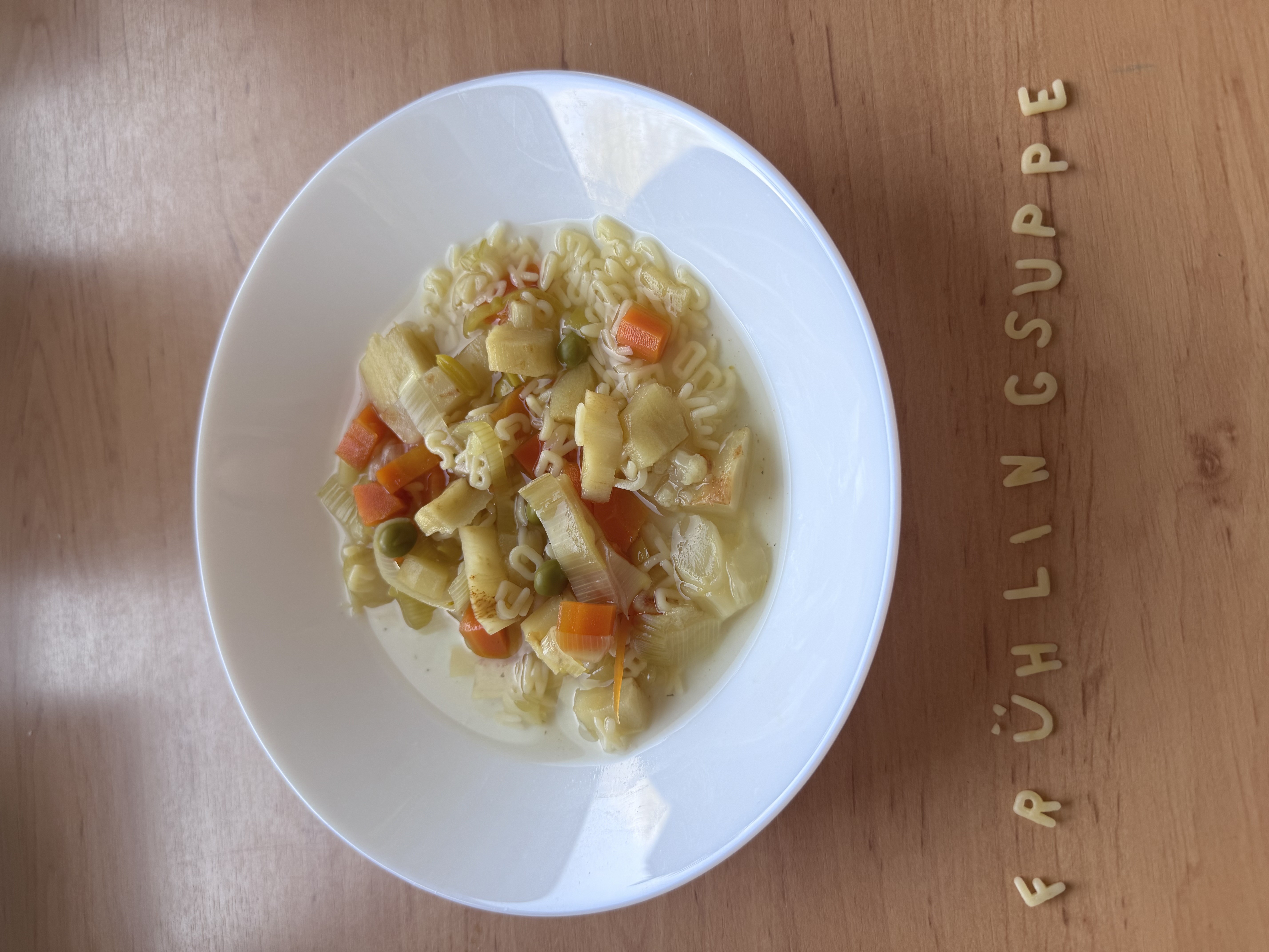 Auf dem Foto steht ein weißer Teller mit einer klaren Gemüsesuppe auf einem Holztisch. In der Suppe sind bunte Gemüsewürfel (z.B. Karotten, Erbsen, Lauch) und Buchstabennudeln zu sehen, und vor dem Teller sind aus Buchstabennudeln die Wörter „FRÜHLING SUPPE“ gelegt.