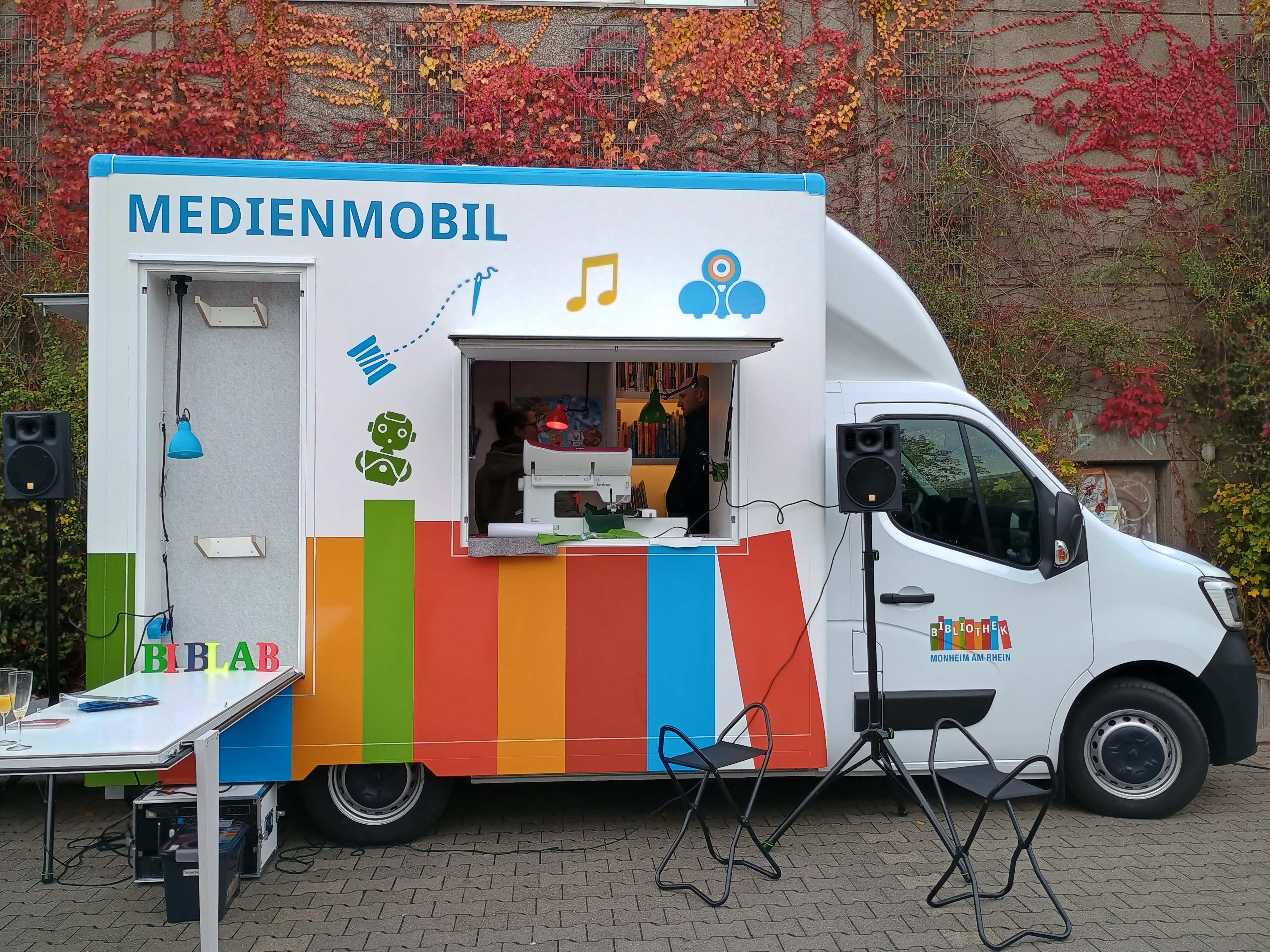 Auf dem Bild ist ein Medienmobil zu sehen, das offensichtlich zu einer Bibliothek gehört (erkennbar am Aufdruck "BIBLAB" und dem Logo der "Stadtbücherei"). Hier sind die Details: Fahrzeug: Es ist ein weißer Kastenwagen mit auffälliger, farbenfroher Gestaltung. Beschriftung: Groß steht "MEDIENMOBIL" auf der Seite. Es gibt auch ein Logo, das die Aufschrift "Stadtbücherei Monheim" trägt. Funktion: Das Mobil hat eine große geöffnete Klappe/Fenster, hinter der man innen jemanden sehen kann. Es scheint ein "BibLab" oder ein mobiler Maker-Space zu sein, denn man sieht eine Nähmaschine in der Öffnung und verschiedene Symbole (Roboter, Musiknote, Kamera/VR-Brille) auf dem Wagen. Umgebung: Das Fahrzeug steht im Freien vor einer Mauer, die dicht mit rot und orange verfärbtem Wein oder Efeu bewachsen ist – deutet auf Herbst hin. Zubehör: Vor dem Mobil stehen Lautsprecher auf Stativen, ein kleiner Tisch mit Kabeln und es gibt zusammenklappbare Stühle.