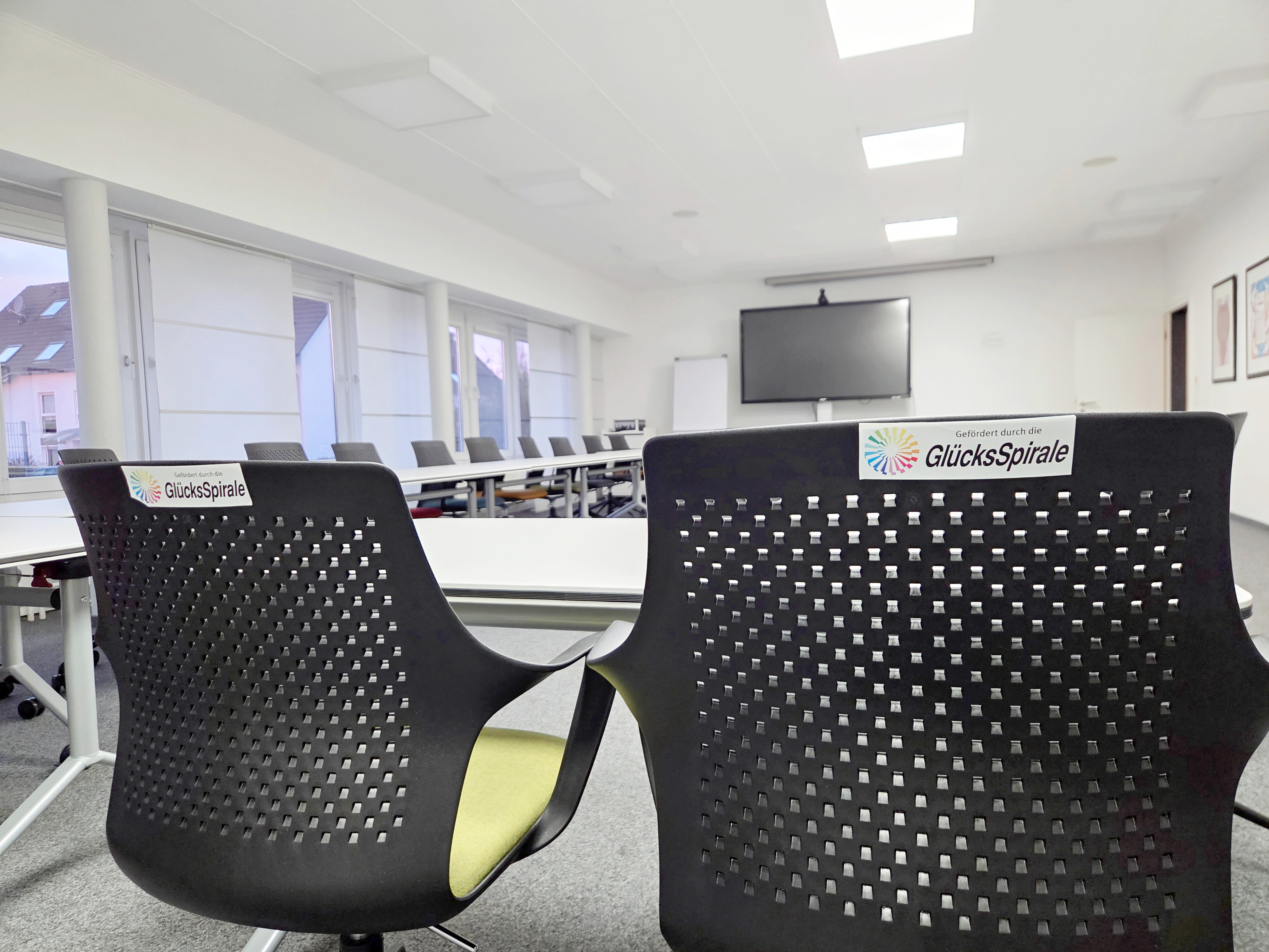 Auf dem Bild ist ein moderner Konferenz- oder Schulungsraum zu sehen, der augenscheinlich neu ausgestattet wurde. Hier sind die Details: Vordergrund: Im Vordergrund sind zwei ergonomisch gestaltete Stühle mit dunklen, perforierten Rückenlehnen zu sehen. Auf beiden Stühlen ist ein Aufkleber mit dem Logo und dem Text "Gefördert durch die GlücksSpirale" angebracht, was auf die Finanzierung der neuen Ausstattung hinweist. Möbel: Im Hintergrund sind weitere Tische und Stühle erkennbar, die in einer U- oder Blockform angeordnet sind. Die Tische scheinen modular und flexibel zu sein, was zu der Beschreibung einer modernen Konferenzausstattung passt. Technik: An der Stirnseite des Raumes ist ein großer Flachbildschirm (vermutlich für Präsentationen oder Videokonferenzen) an der Wand montiert. Raumgestaltung: Der Raum wirkt hell und aufgeräumt mit einer abgehängten Decke, in die quadratische LED-Leuchten eingelassen sind. Die Fensterfront ist mit hellen Rollos oder Jalousien versehen. Gesamteindruck: Das Bild vermittelt den Eindruck eines professionellen, zeitgemäßen Tagungsortes, der für Schulungen, Besprechungen oder Konferenzen eingerichtet wurde.