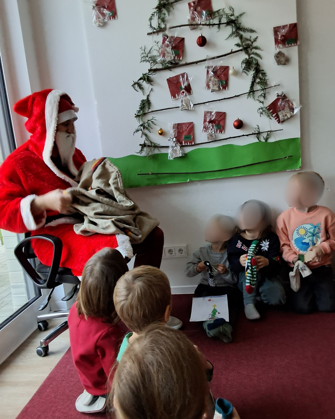 Auf dem Bild ist eine Szene zu sehen, die an die Weihnachtszeit erinnert. Hier sind die Details: Der Nikolaus/Weihnachtsmann: Eine Person in einem roten Kostüm mit weißem Bart und Kapuze (die Figur des Nikolaus oder Weihnachtsmannes) sitzt auf einem Bürostuhl. Die Person scheint gerade einen Sack oder eine Tasche zu öffnen oder etwas daraus hervorzuholen. Kinder: Mehrere Kleinkinder sitzen auf dem Boden vor der als Nikolaus verkleideten Person. Einige schauen zu, während sie warten. Die Gesichter der Kinder sind unkenntlich gemacht (verpixelt/unscharf). Hintergrund und Dekoration: An der Wand hängt eine kreative, stilisierte Weihnachtsbaum-Dekoration. Sie besteht aus grünen Zweigen, die in Form eines Baumes angebracht sind. Daran hängen kleine Päckchen (möglicherweise Adventskalendertüten oder Geschenke) und einige rote Weihnachtskugeln. Der untere Teil der Dekoration ist eine grüne Bahn, die wahrscheinlich den Boden oder eine Wiese darstellen soll.
