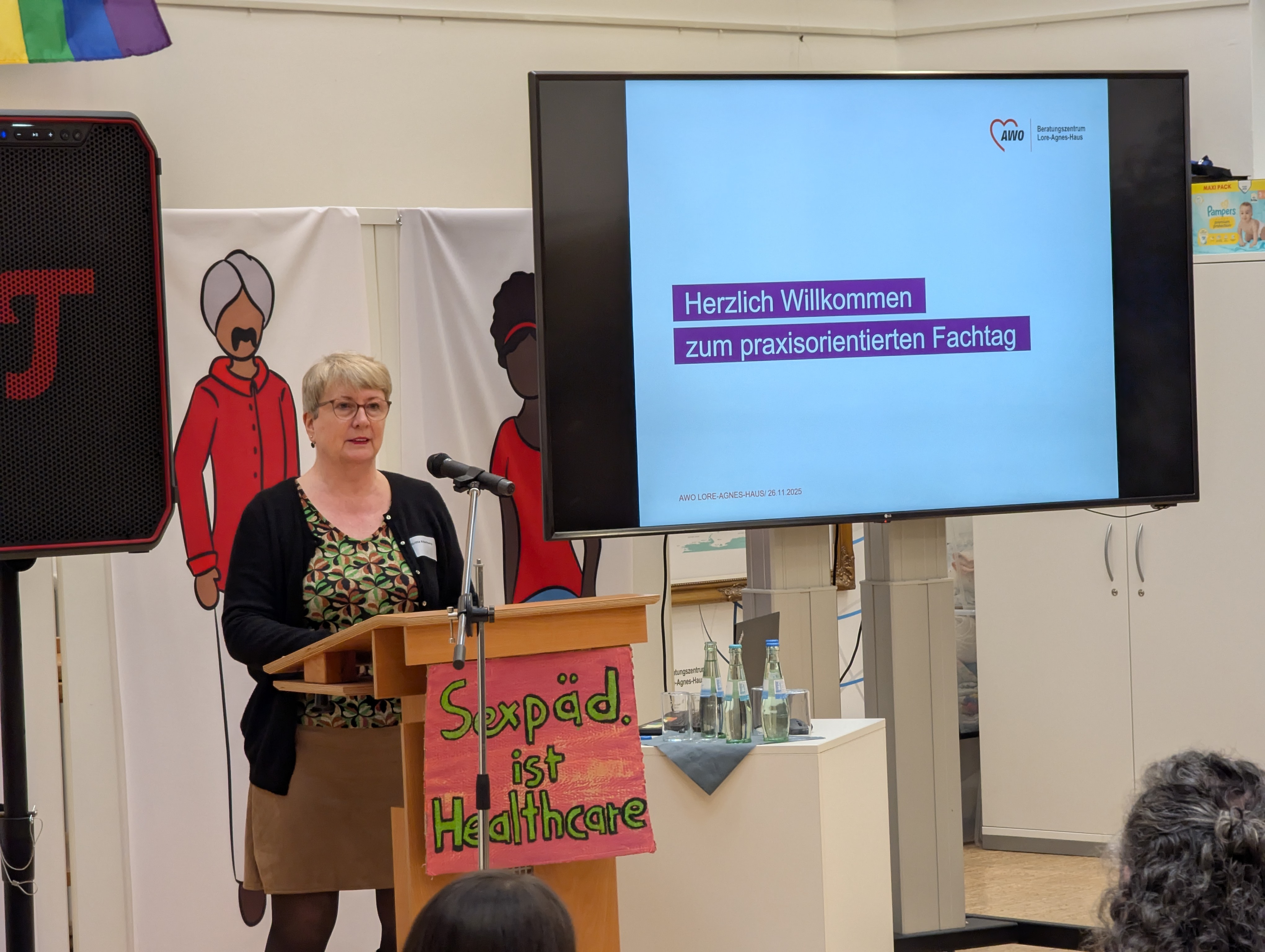 Auf dem Bild ist eine Veranstaltung oder Konferenz zu sehen. Hier sind die Details: Vordergrund: Eine Frau steht an einem Holzpult und spricht in ein Mikrofon. Sie trägt eine Brille, ein gemustertes Oberteil und eine dunkle Strickjacke. Pult-Dekoration: Am Pult hängt ein Schild, auf dem in bunter Schrift steht: "Sexpäd. ist Healthcare" (was wahrscheinlich "Sexualpädagogik ist Gesundheitsfürsorge" bedeutet). Hintergrund (Leinwand/Bildschirm): Ein großer Bildschirm zeigt eine Präsentation mit dem Text: "Herzlich Willkommen zum praxisorientierten Fachtag" Darunter steht das Logo der AWO (Arbeiterwohlfahrt) und der Text "Beratungszentrum Lippe/OWL" sowie das Datum "AWO JULE AUBI | 26.11.2025". Weitere Hintergründelemente: Links hinter der Rednerin stehen Banner mit stilisierten Illustrationen von Menschen. Ganz links ist ein Lautsprecher zu erkennen. Rechts neben dem Pult steht ein kleiner Tisch mit Wasserflaschen und Gläsern.