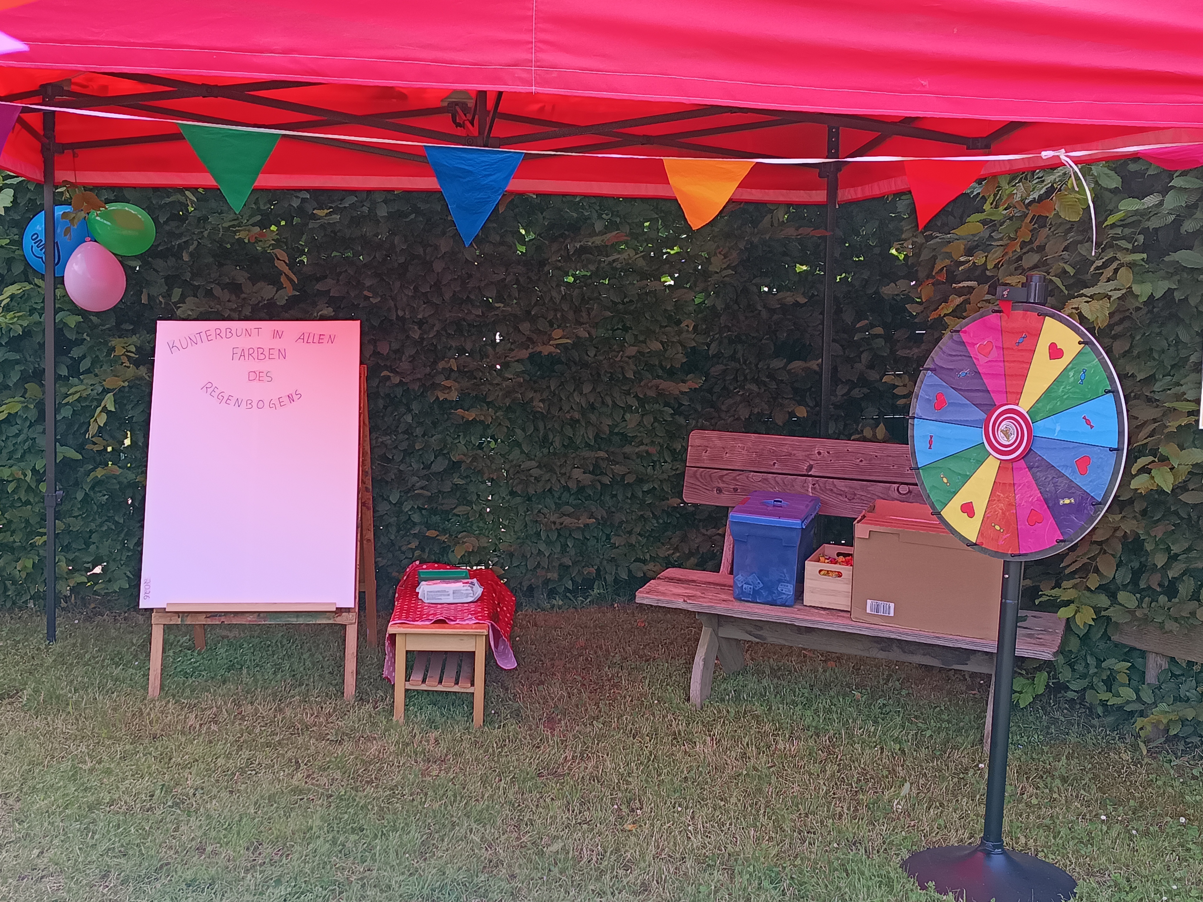 Auf dem Bild ist ein kunterbunt gestalteter Aktionsstand auf einer Wiese unter einem roten Pavillon zu sehen. Elemente des Standes Links steht eine große weiße Tafel auf einem Holzständer, oben mit dem Schriftzug „Kunterbunt in allen Farben des Regenbogens“. Daneben steht ein kleiner Tisch mit rot‑weiß karierter Decke und Materialien darauf. Umgebung und Deko Rechts im Bild befindet sich ein bunt bemaltes Glücksrad mit Herzmotiven vor einer Holzbank, auf der Kisten und Boxen stehen. Unter dem Pavillon hängen bunte Wimpelketten und Ballons, im Hintergrund ist eine dichte grüne Hecke zu erkennen